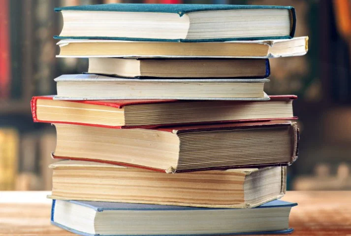 Stack of several hardcover and paperback books on a wooden surface with blurred bookshelves in the background.