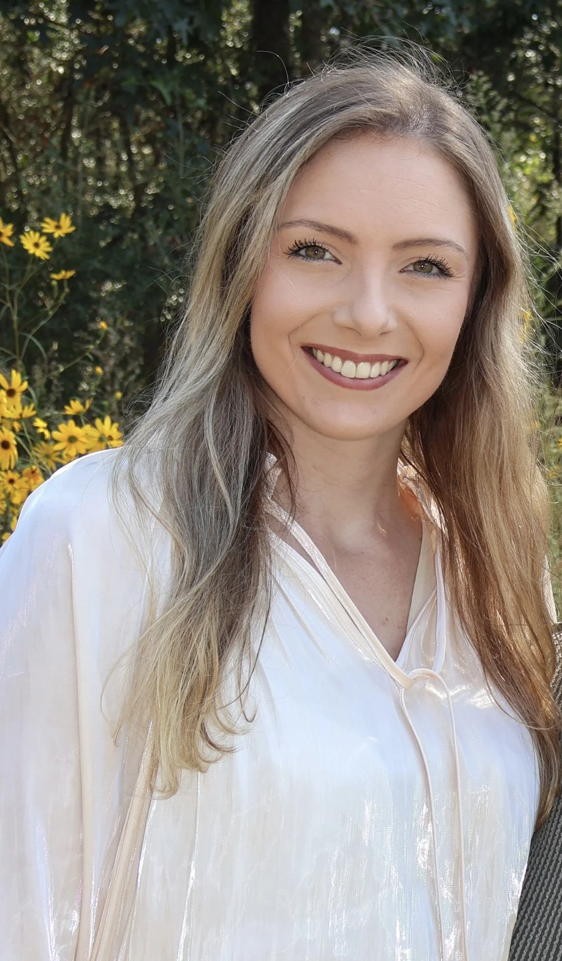 A young woman with long, wavy blonde hair and hazel eyes smiling outdoors near yellow flowers and green foliage.