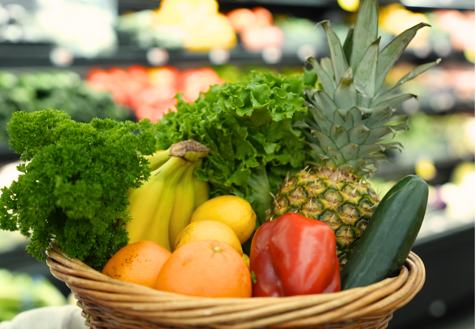 A basket of fresh fruits and vegetables including pineapple, cucumber, red bell pepper, citrus fruits, bananas, lettuce, and parsley on a grocery store shelf.