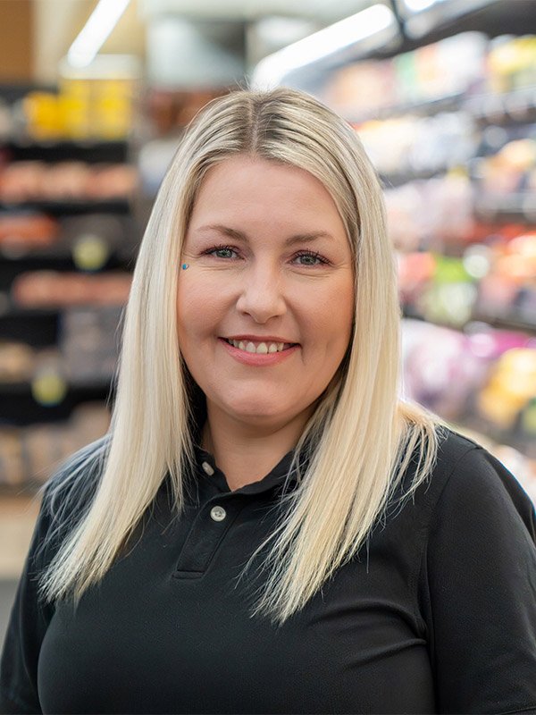 A woman with long blonde hair and a small blue beauty mark below her right eye, smiling in a grocery store aisle.