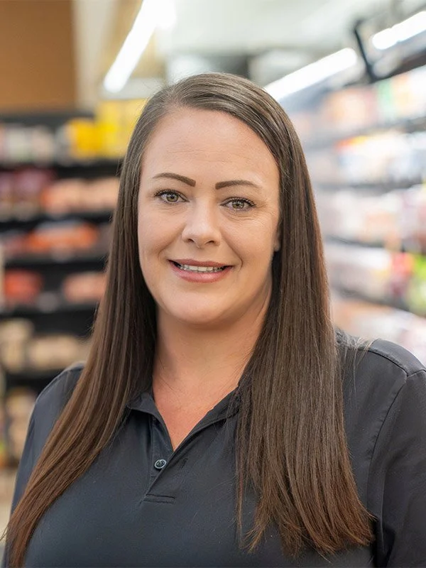 A woman with long brown hair and a black shirt smiling in a grocery store aisle.