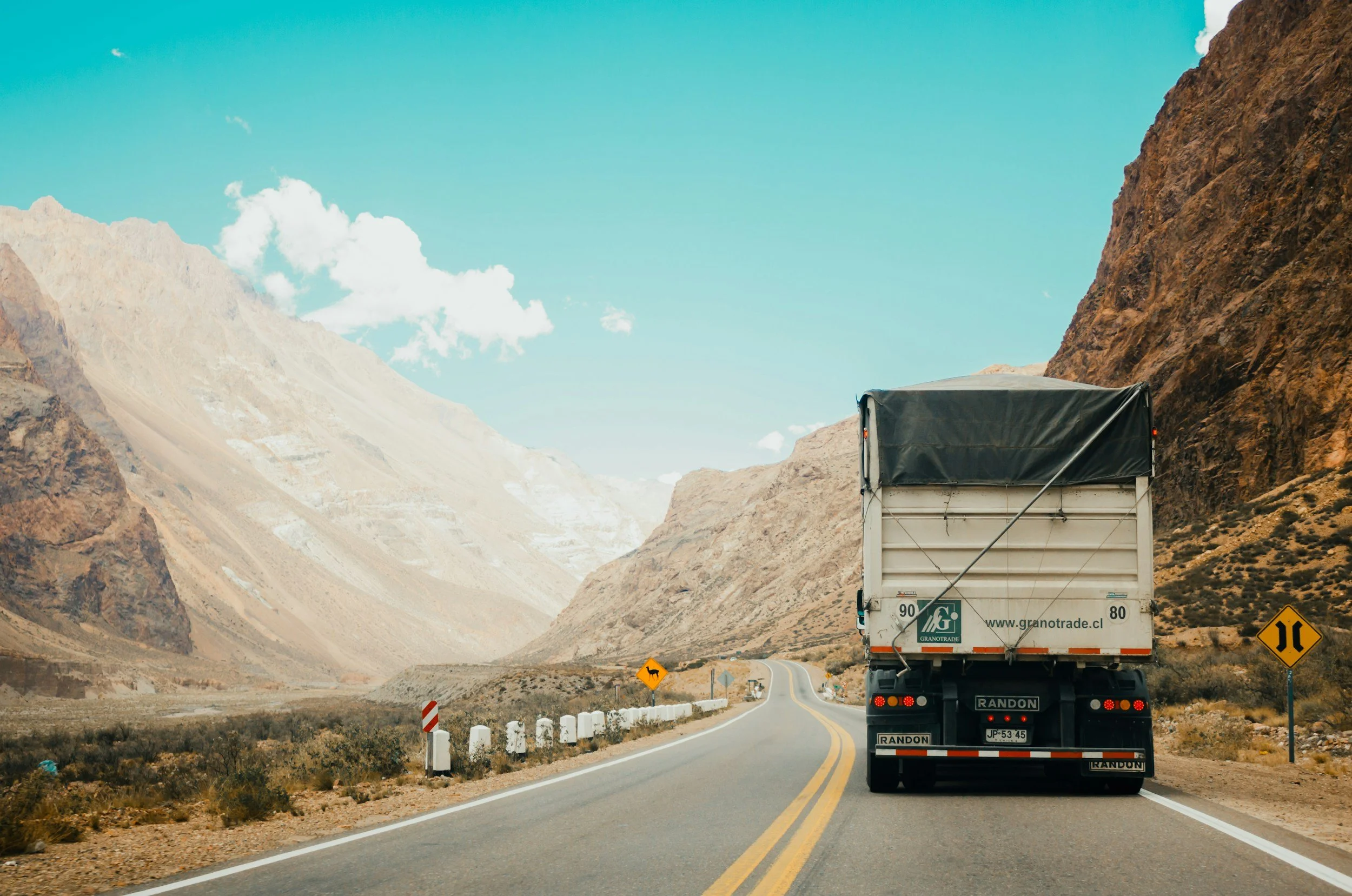 A truck driving on a winding mountain road with rocky cliffs on both sides under a blue sky with scattered clouds.