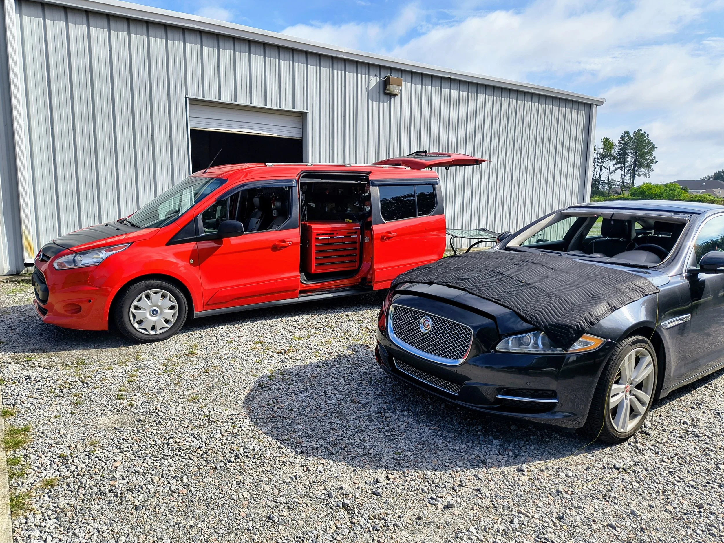 A red utility van and a black luxury car with a quilted cover on the hood parked outside a metal building on a gravel lot under a partly cloudy sky.
