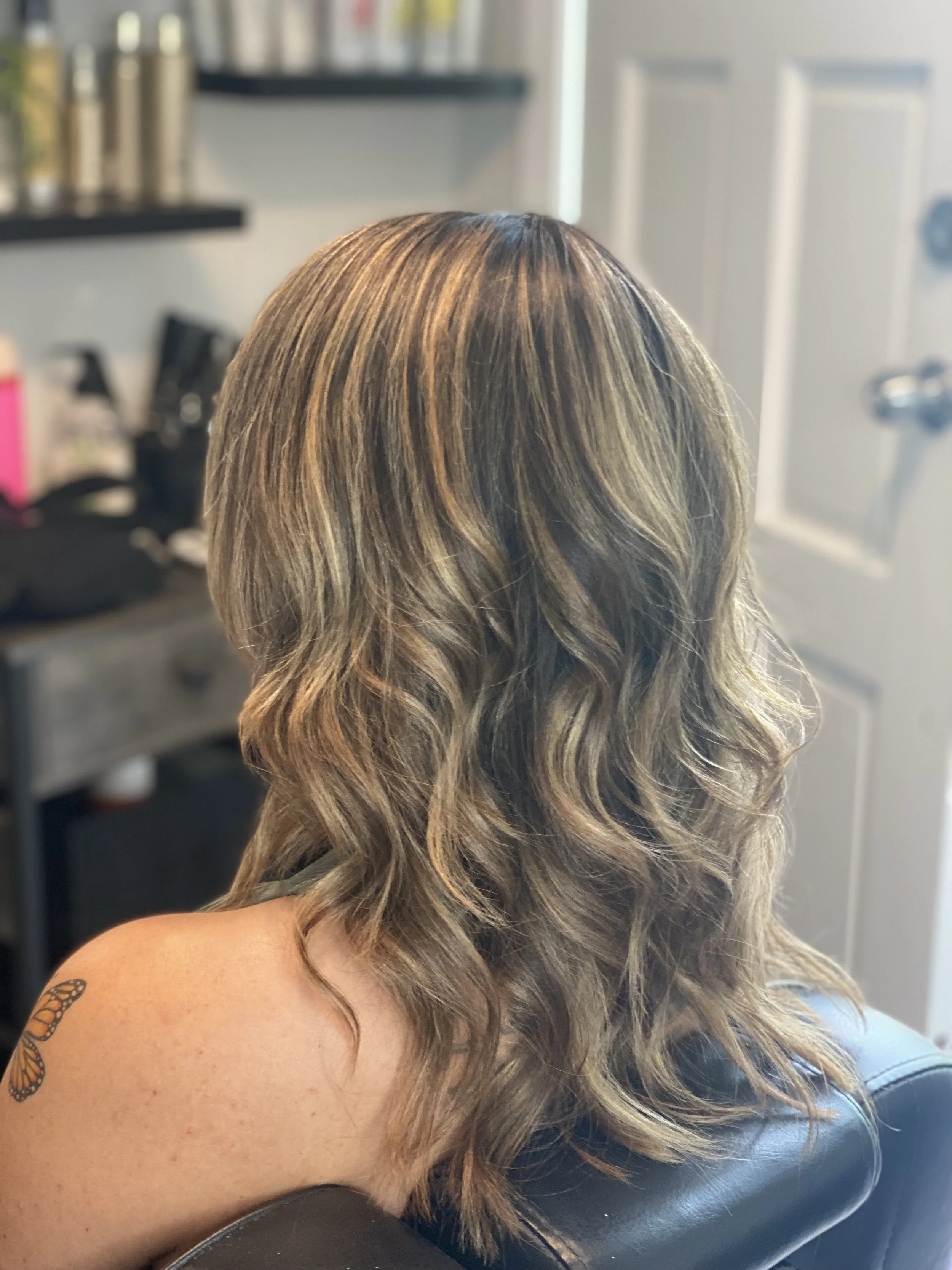 Back of a woman's head showing shoulder-length, wavy, multi-toned blonde hair with highlights, sitting in a salon chair.