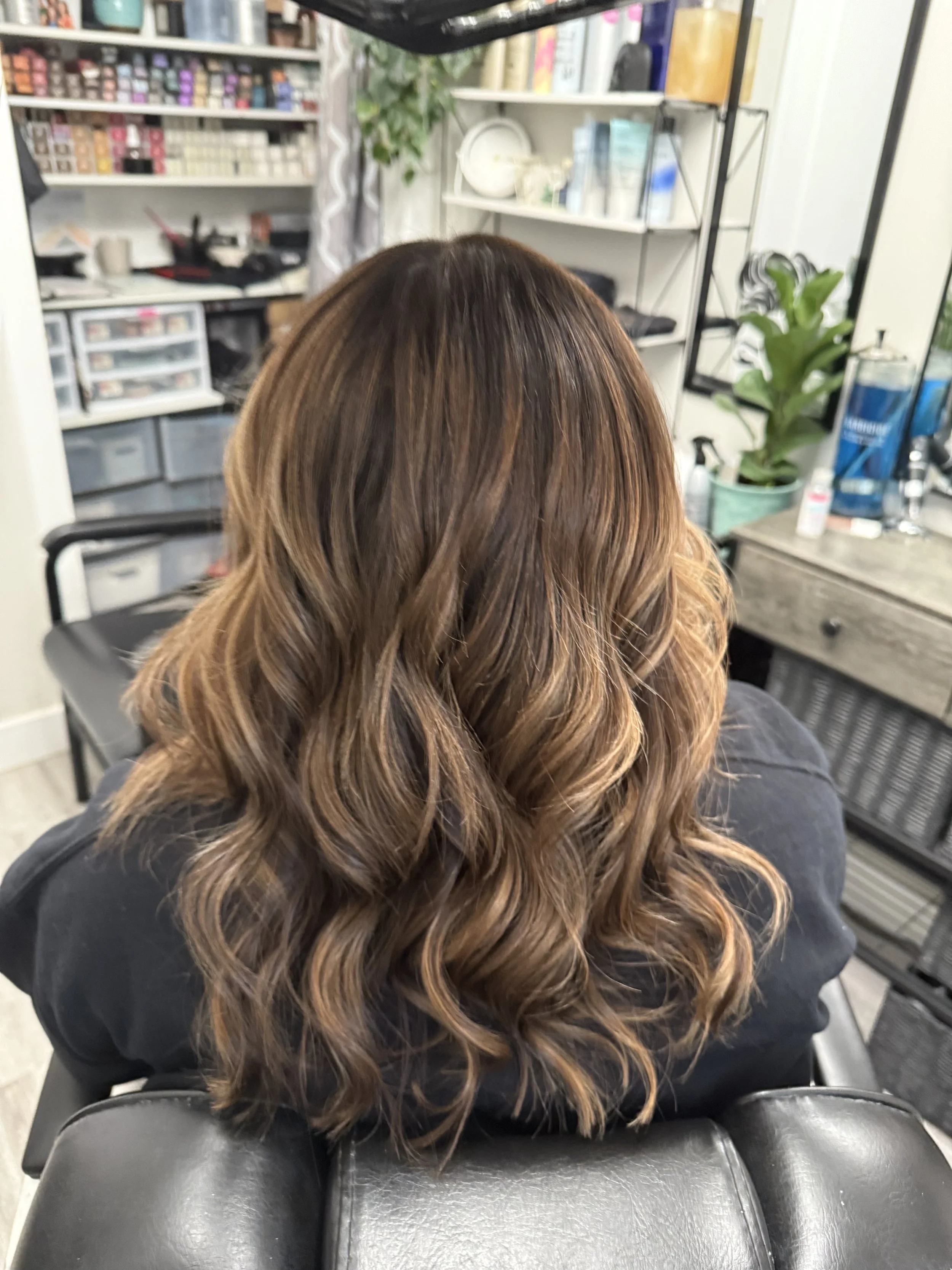 Back view of a woman with wavy, brown hair in a salon chair.