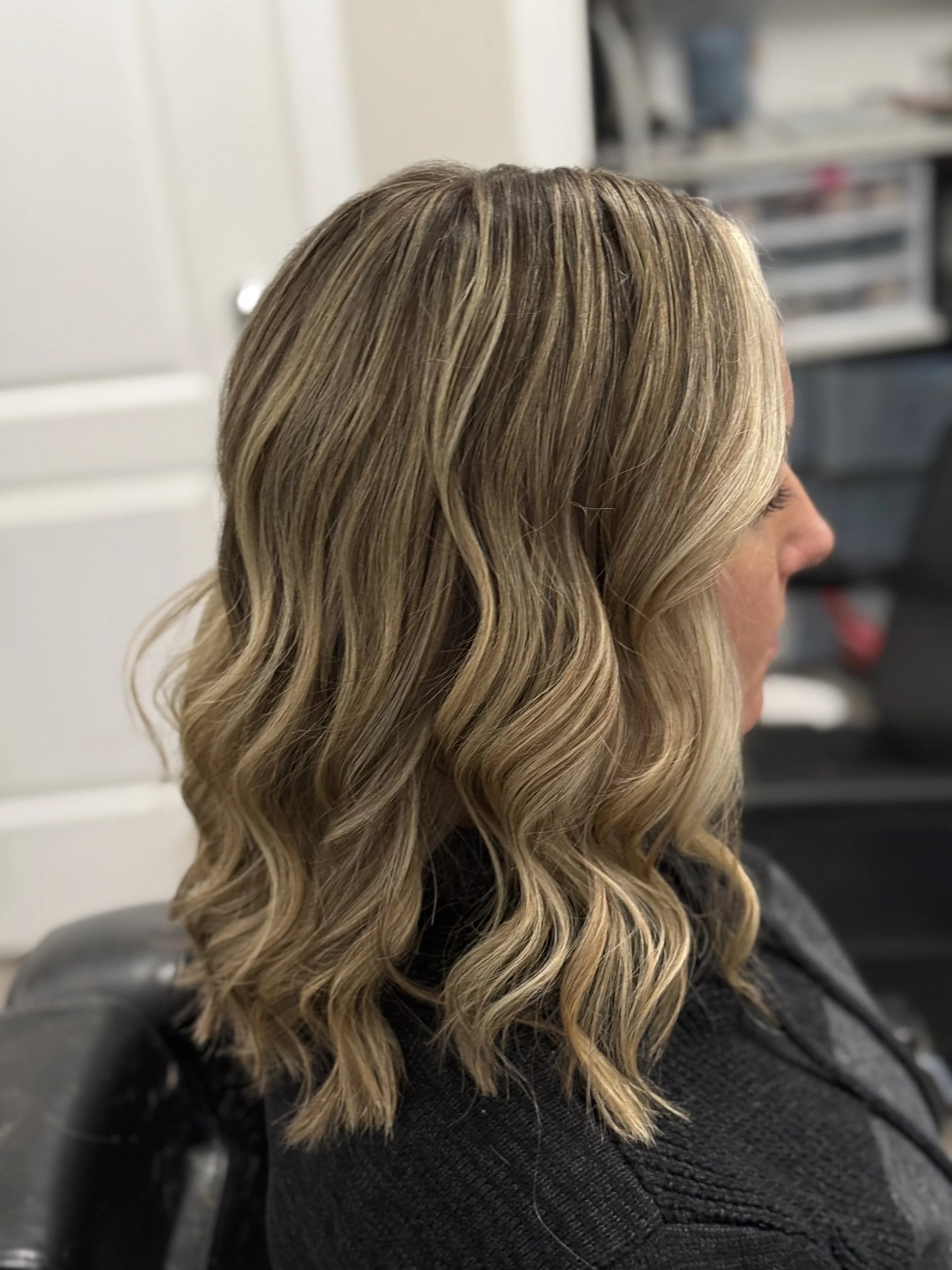 Side view of a woman with blonde, wavy shoulder-length hair sitting in a salon chair.
