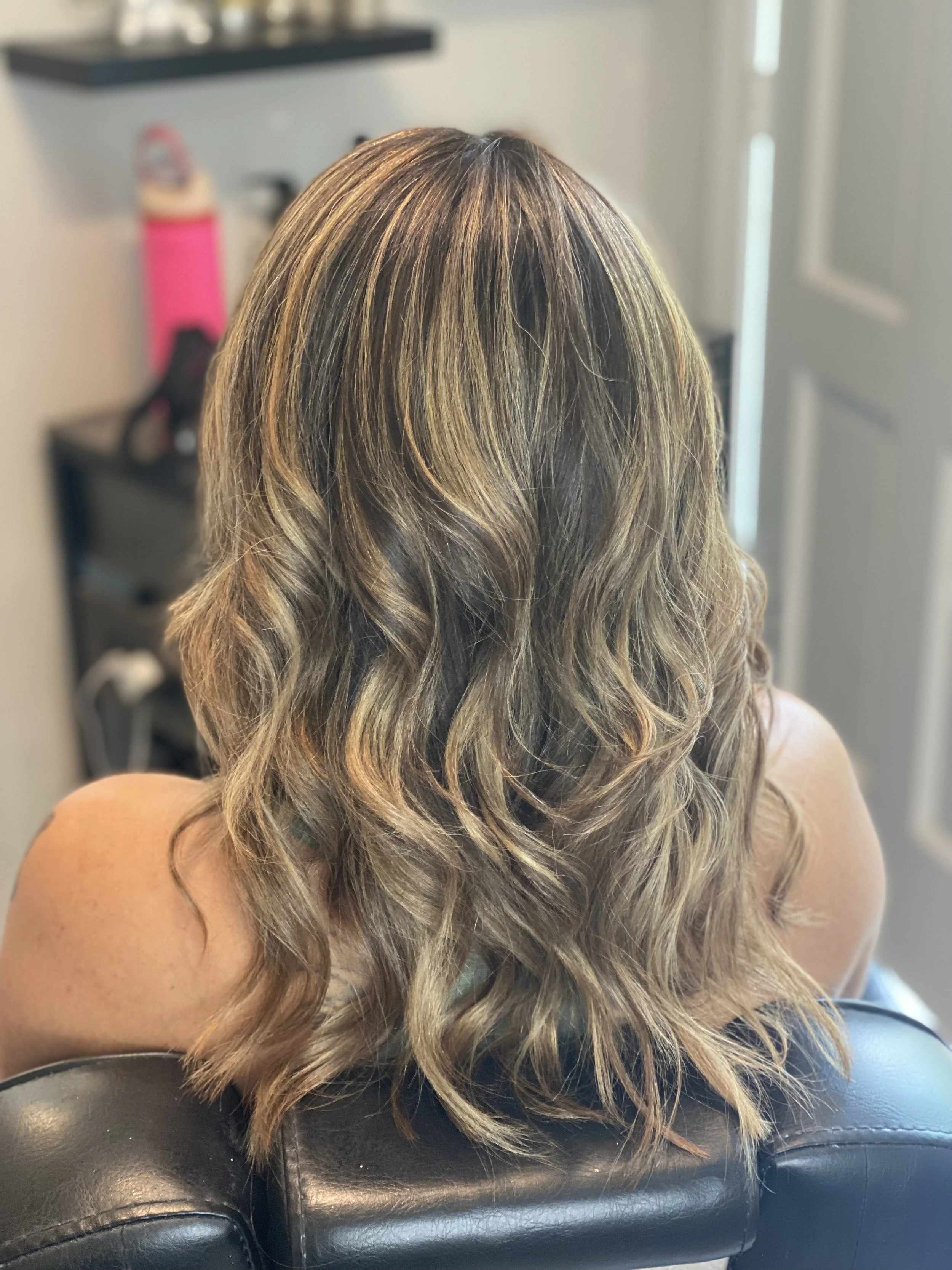 Back view of a woman with shoulder-length, wavy, blonde hair sitting in a salon chair.