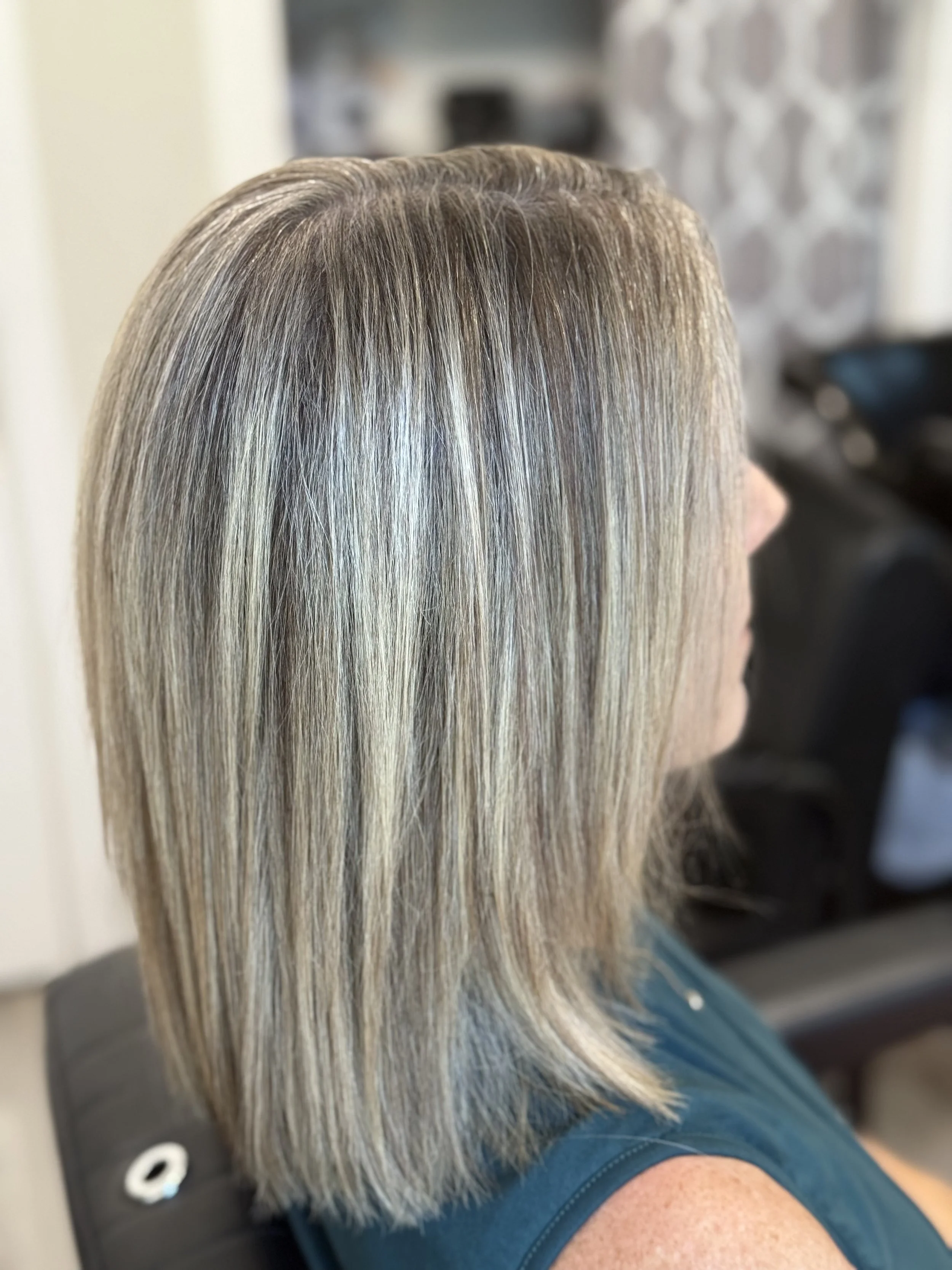 Side view of a woman with shoulder-length blonde hair with highlights, sitting in a salon chair.