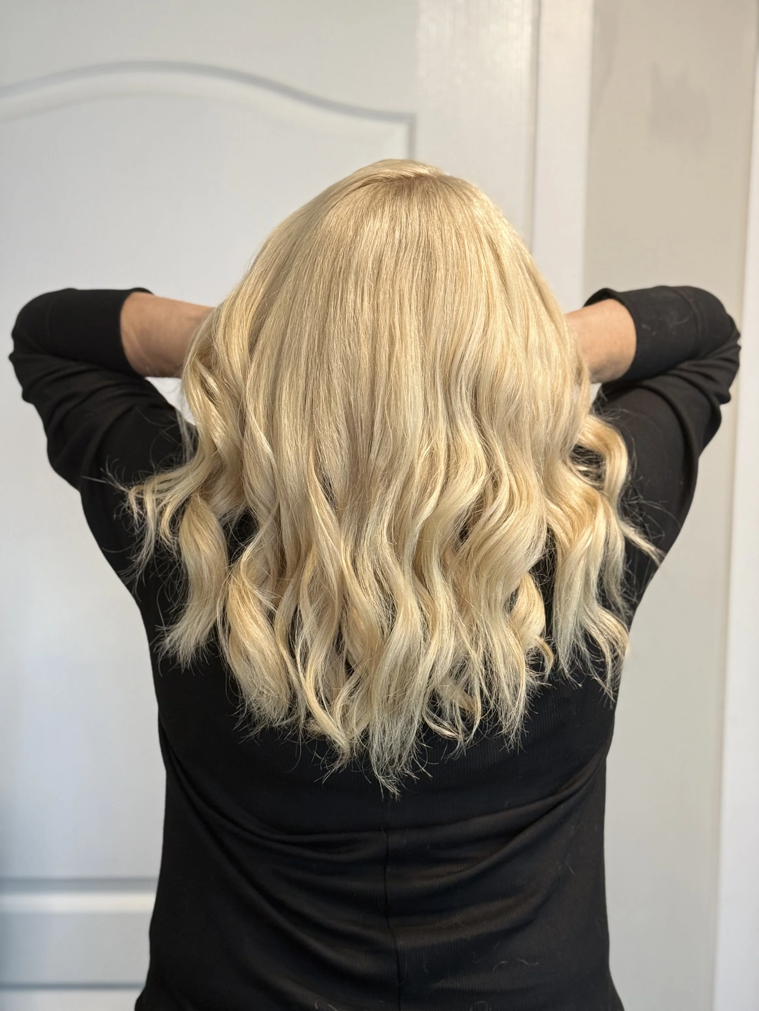 Back view of a woman with long, wavy blonde hair wearing a black top, standing in front of a light-colored door.