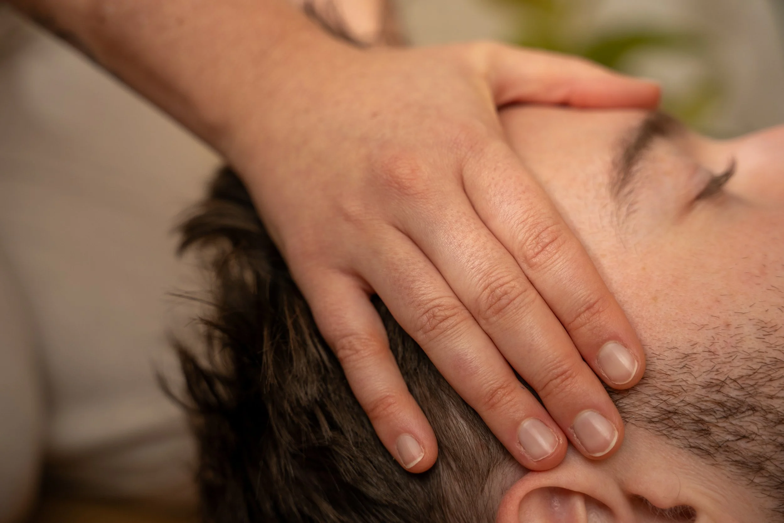 A close-up of a person's head with short dark hair, receiving a massage with a hand placed on their scalp.