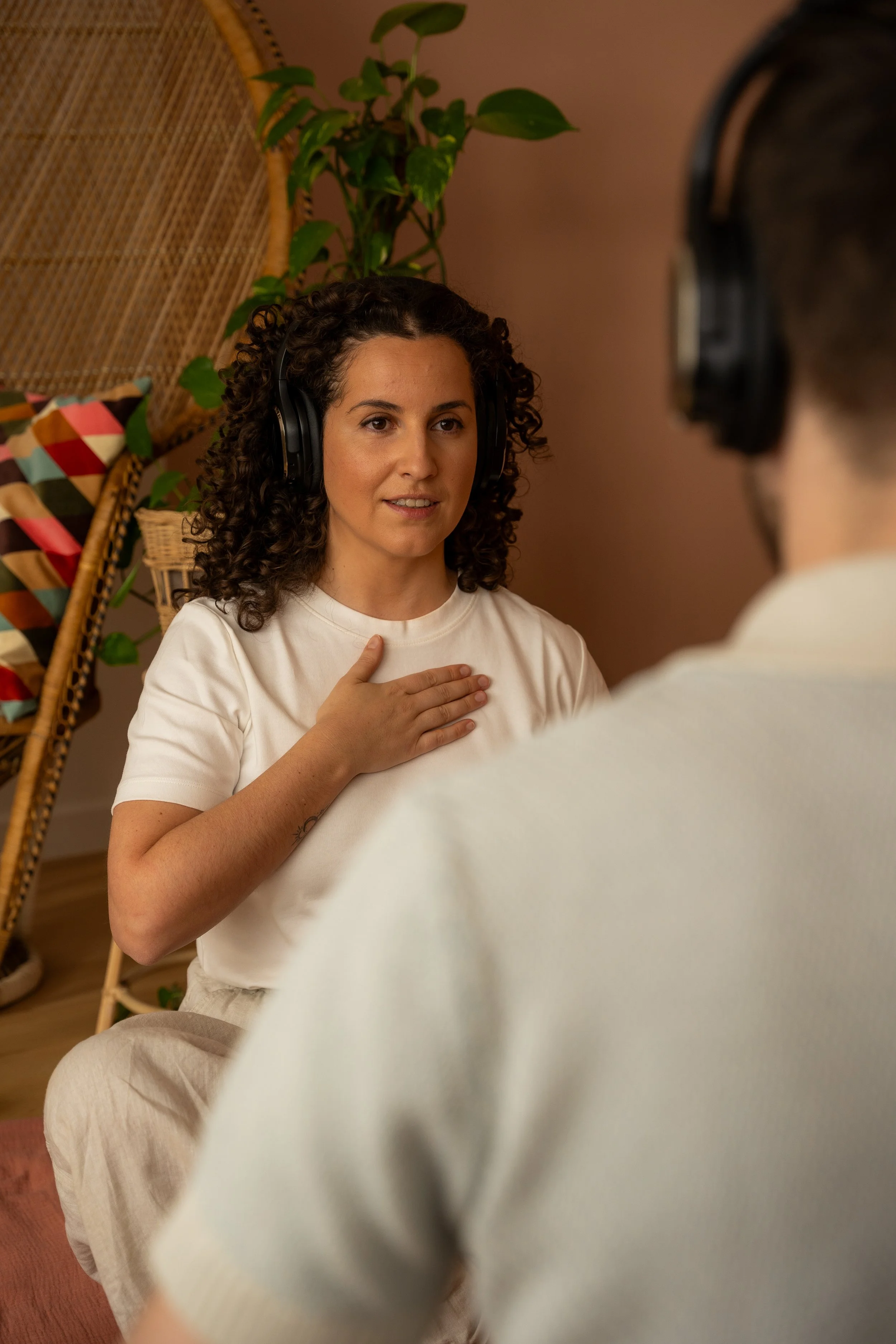 A woman sits on the floor with her hand over her heart, wearing headphones, and appears to be speaking or singing during a therapy or counseling session with a man, who is partially visible from behind, also wearing headphones.