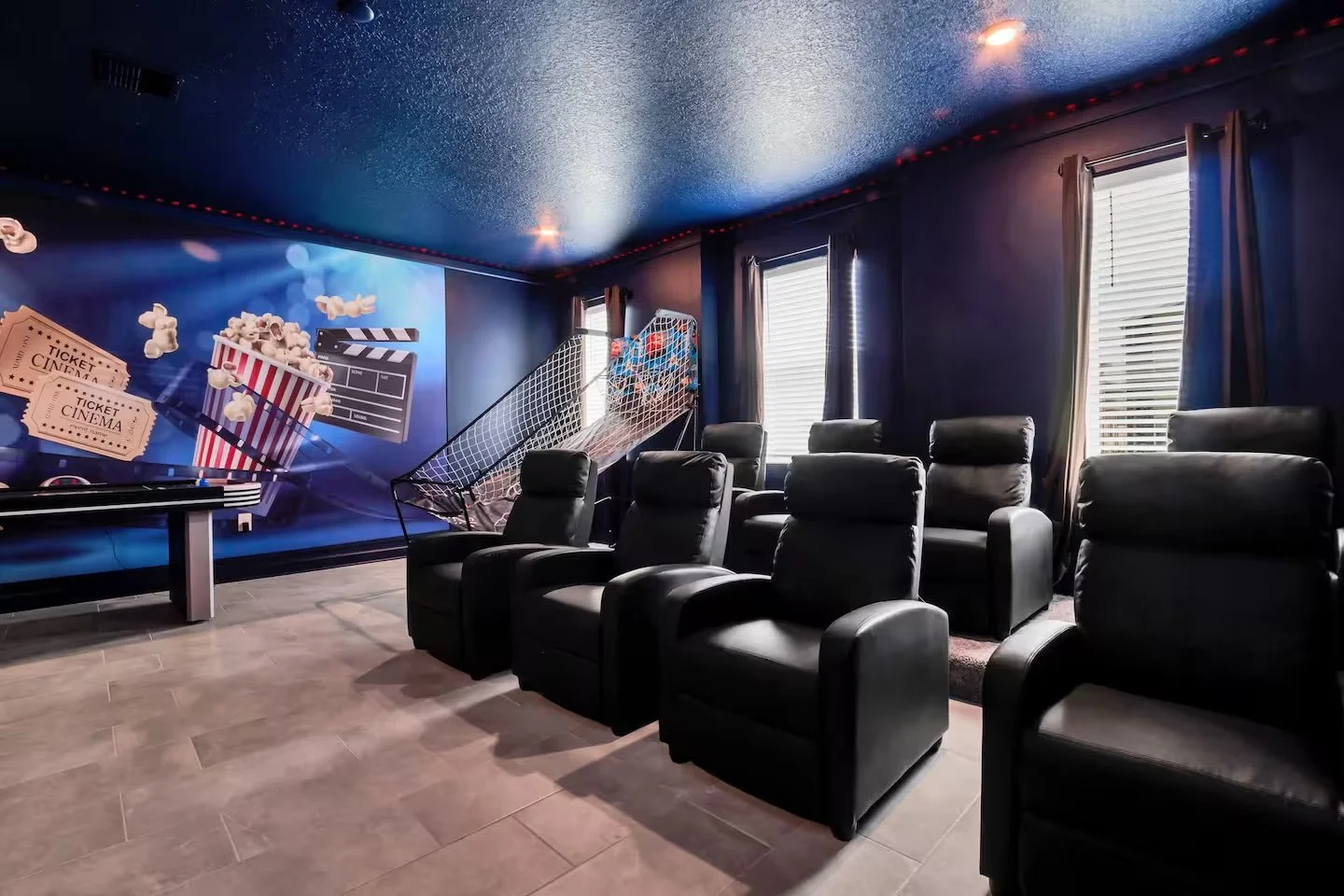 Room with two rows of black leather recliner chairs facing a wall with movie-themed mural, three windows with blinds and curtains, and a popcorn machine. Vacation rental movie room in Storey Lake, Kissimmee.