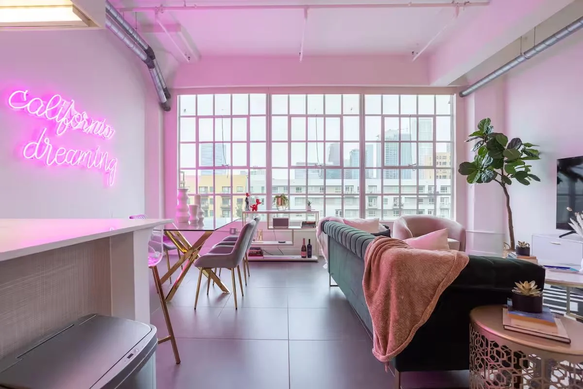 Modern living room with pink neon sign saying 'California dreaming,' large windows with city skyline view, green sofa with blanket, coffee table with books and plants, and a TV on the right side.