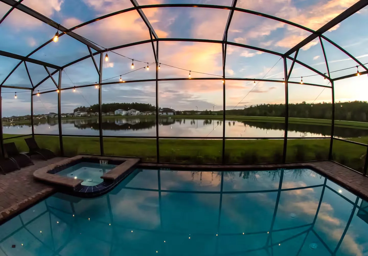 View of a screened porch with a hot tub and swimming pool, overlooking a lake during sunset with colorful clouds and a calm water reflection. Beautiful lakefront vacation home rental in Storey Lake, Kissimmee, in Orlando Florida