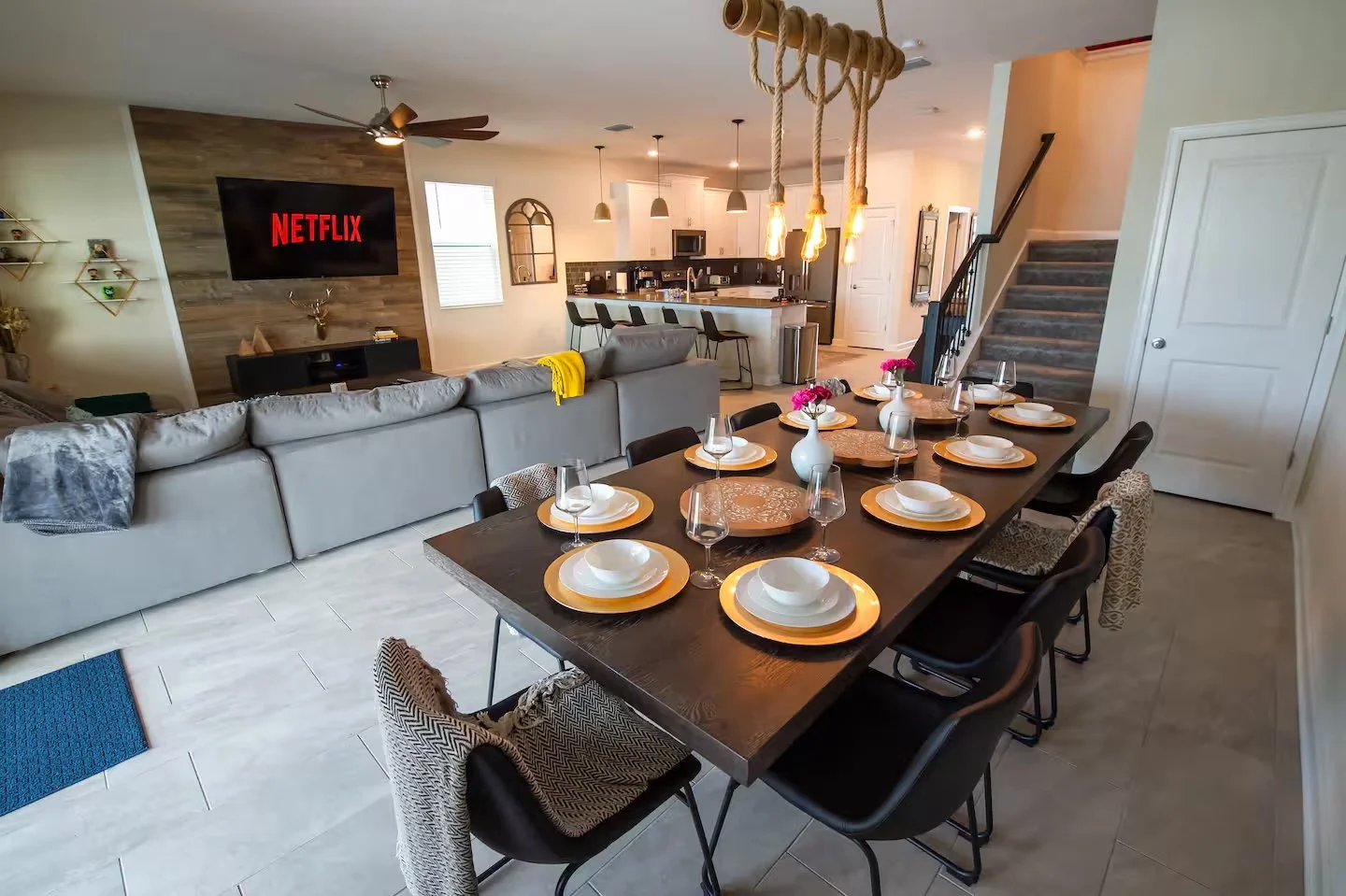 Open-concept living and dining area with a large dark dining table set with white and gold dinnerware, a decorated living area with a TV displaying Netflix, and a kitchen with a breakfast bar and hanging light fixtures.