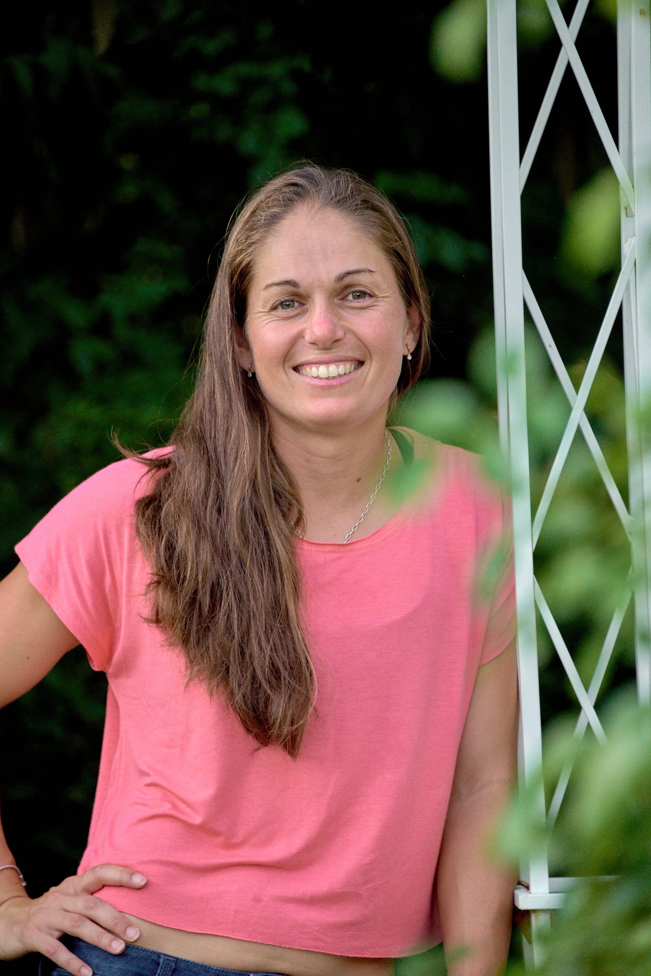 A woman with long brown hair wearing a pink t-shirt, standing outdoors next to a white lattice structure, smiling with a background of green foliage.