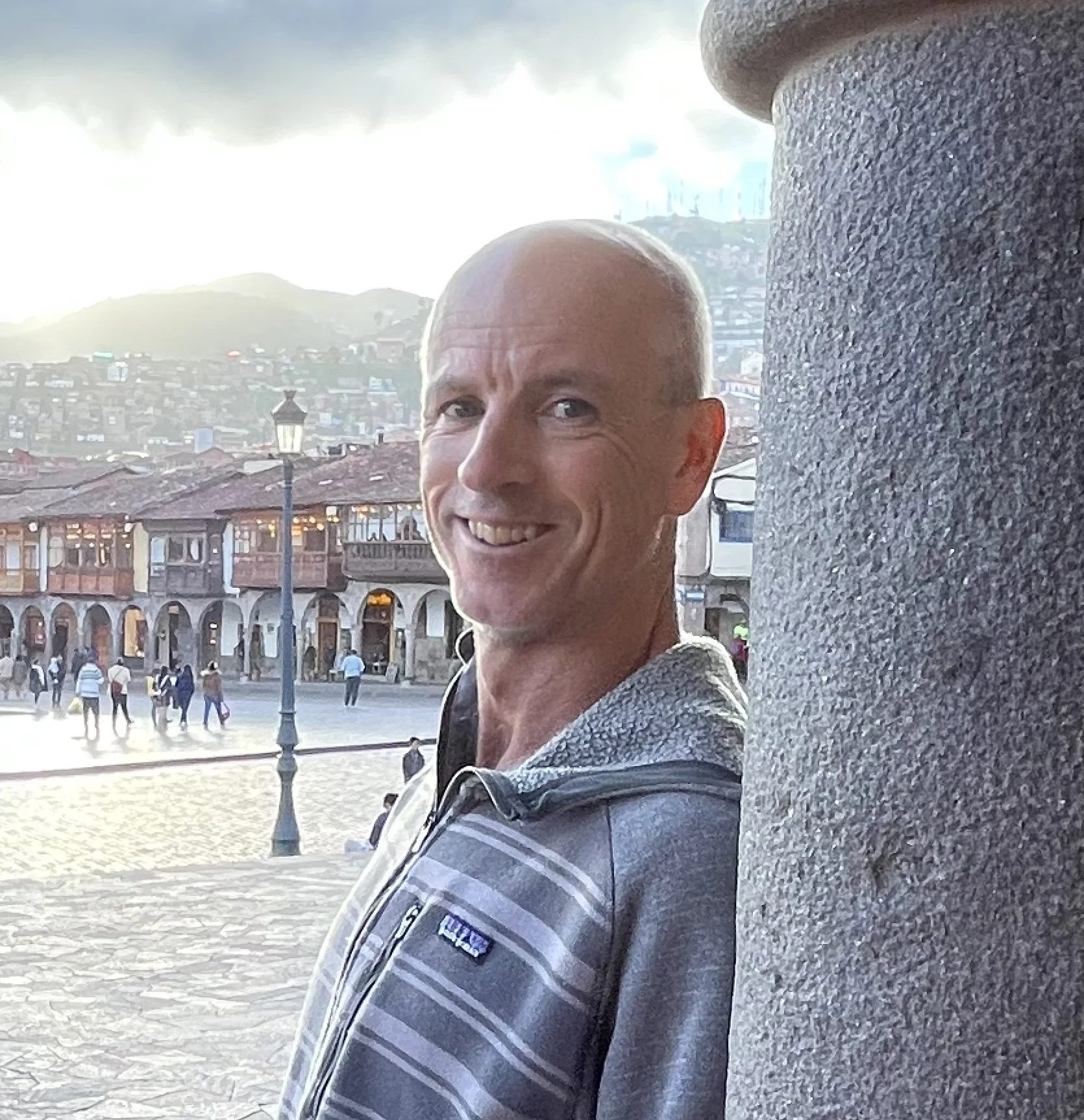 Smiling man with a bald head and a striped jacket stands near a stone pillar in a public square with people and buildings in the background.