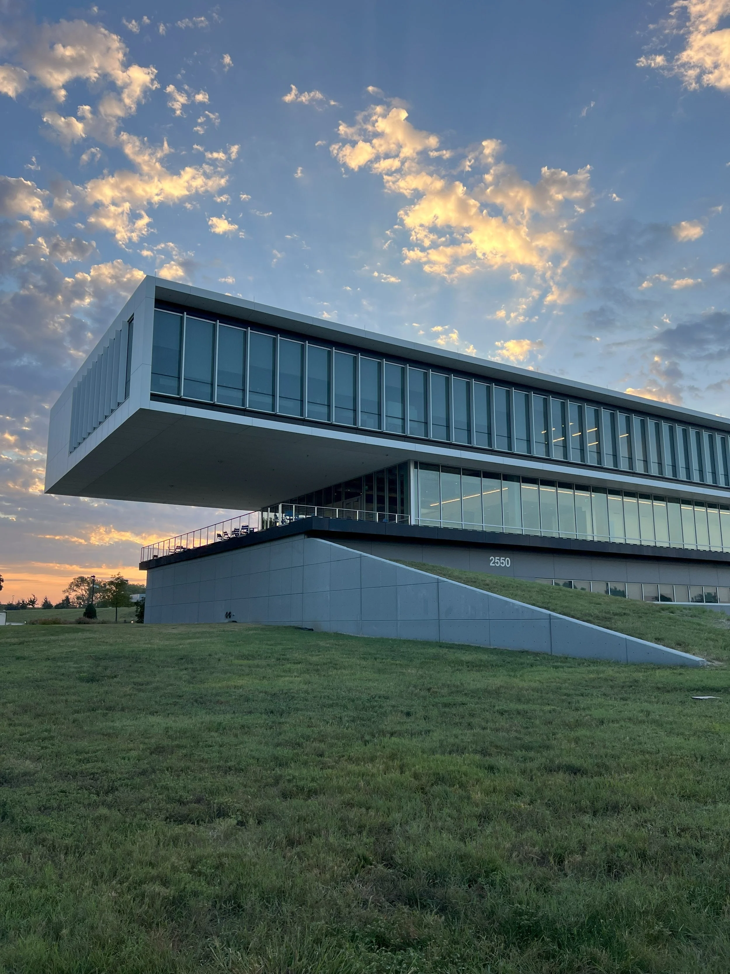 Modern building with large glass windows, cantilevered upper floor, surrounded by green grass and a partly cloudy sky at sunset.