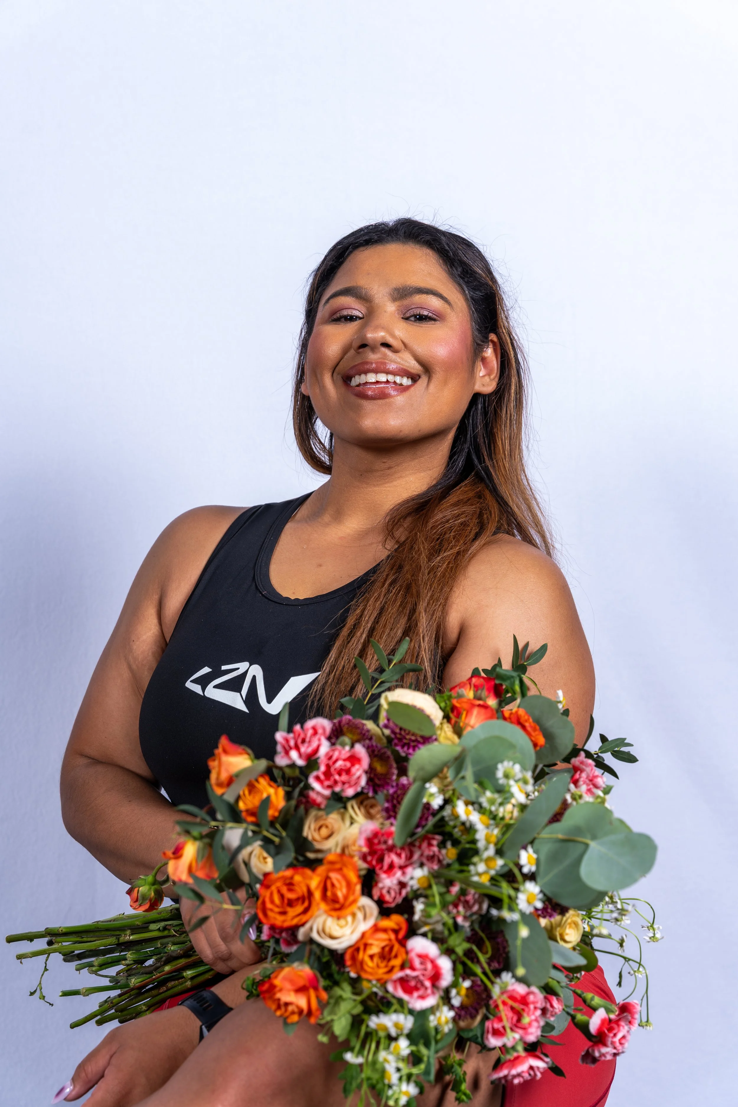 A woman holding a large bouquet of colorful flowers, smiling, wearing a black tank top with a logo, standing against a plain white background.