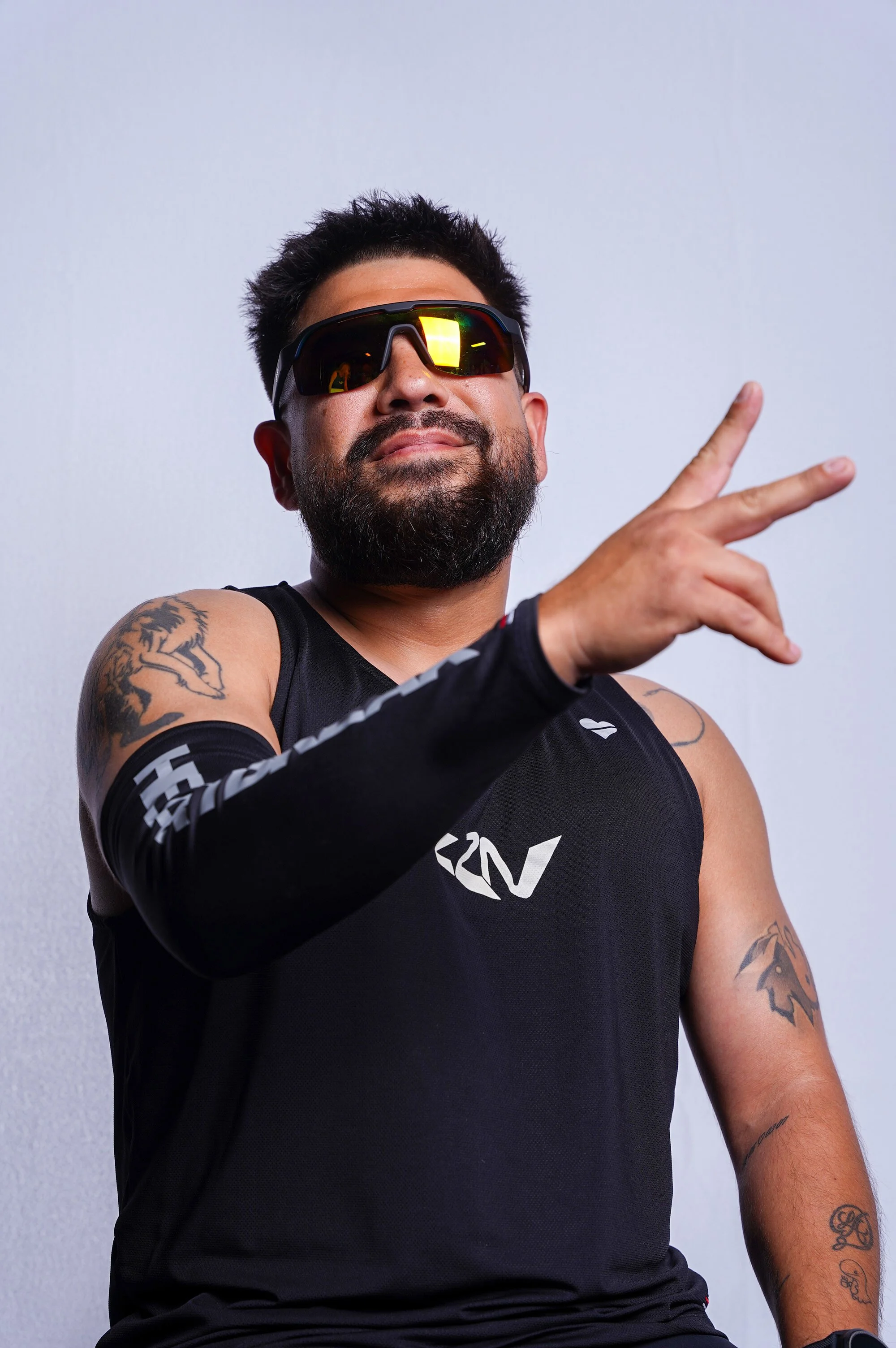 A man with a beard and tattoos making a peace sign gesture while wearing sunglasses and a black sleeveless athletic shirt.