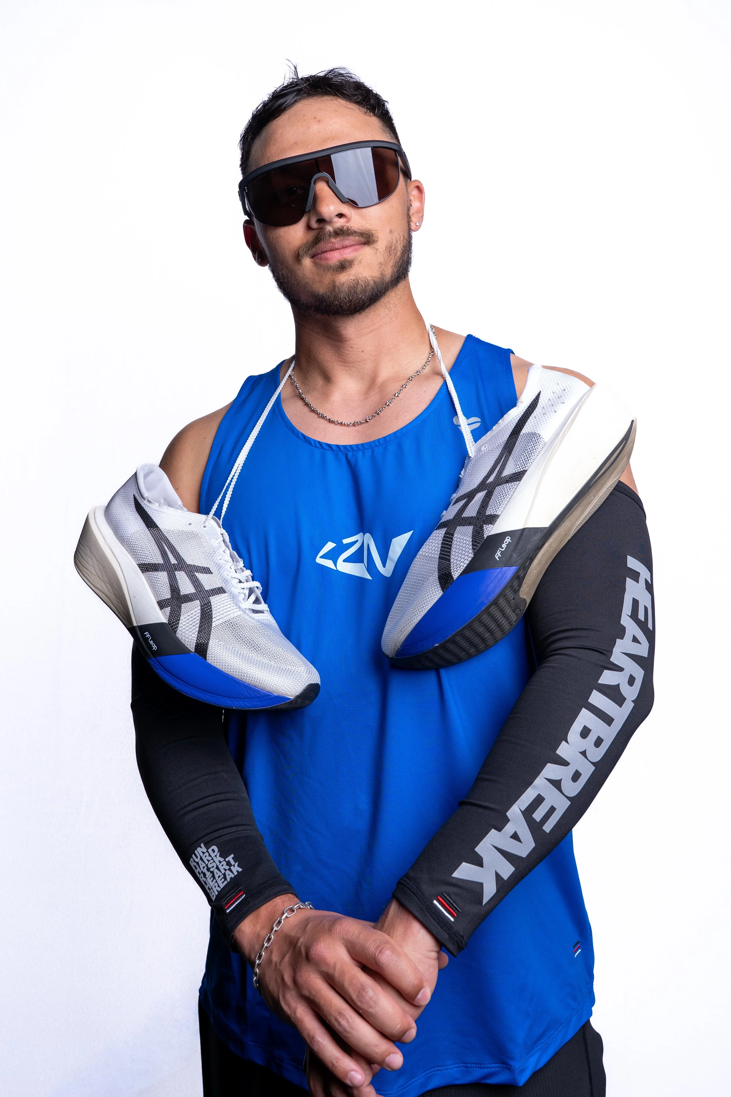 A fit man wearing sunglasses, a blue athletic tank top, and black arm sleeves with the word 'HEARTBREAK' on one sleeve, holding a pair of running shoes with white, black, and blue accents draped over his shoulders, standing against a plain white background.