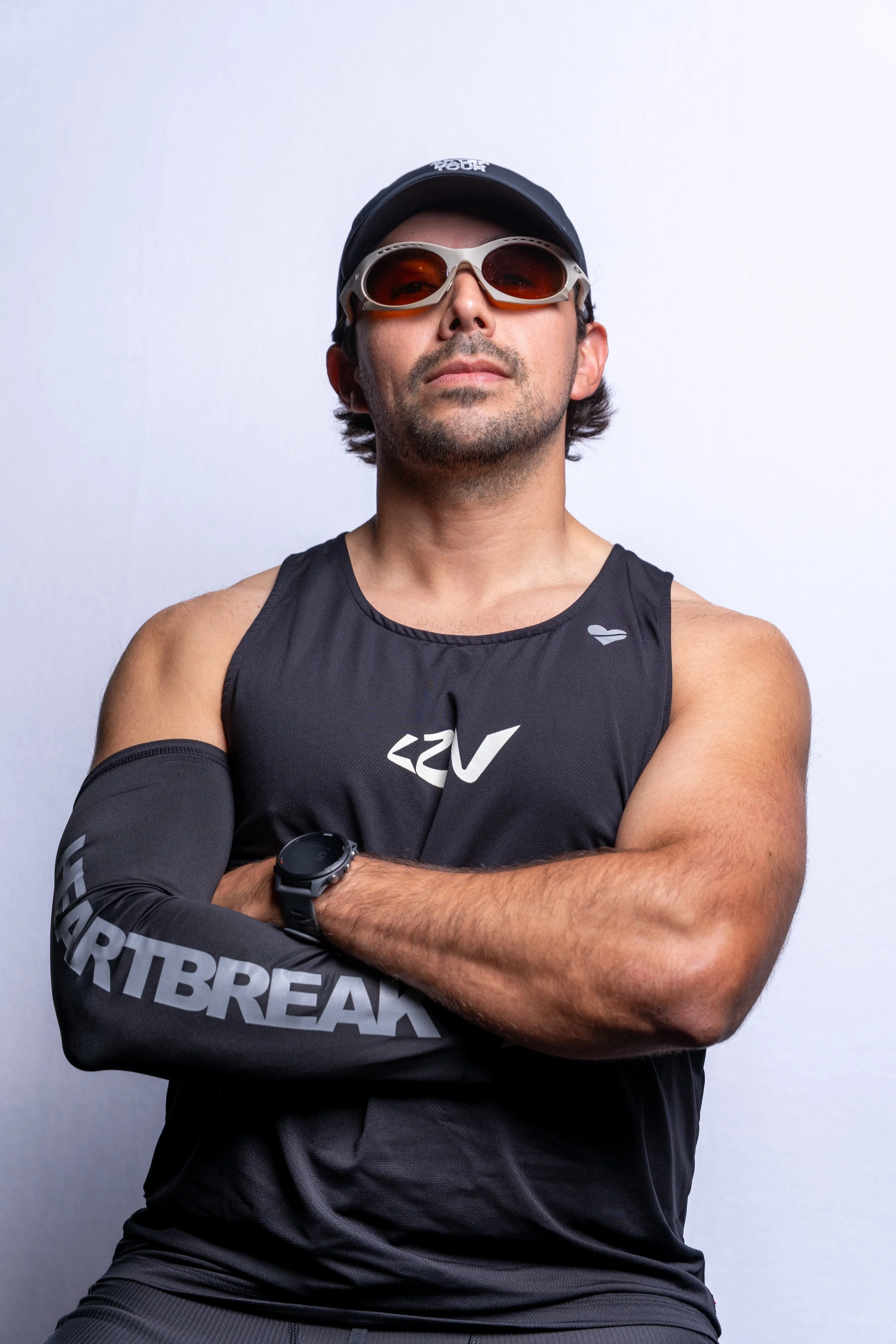 A confident man wearing sunglasses, a black cap, a sports arm sleeve, a sleeveless athletic shirt, and a watch, standing with arms crossed against a plain gray background.