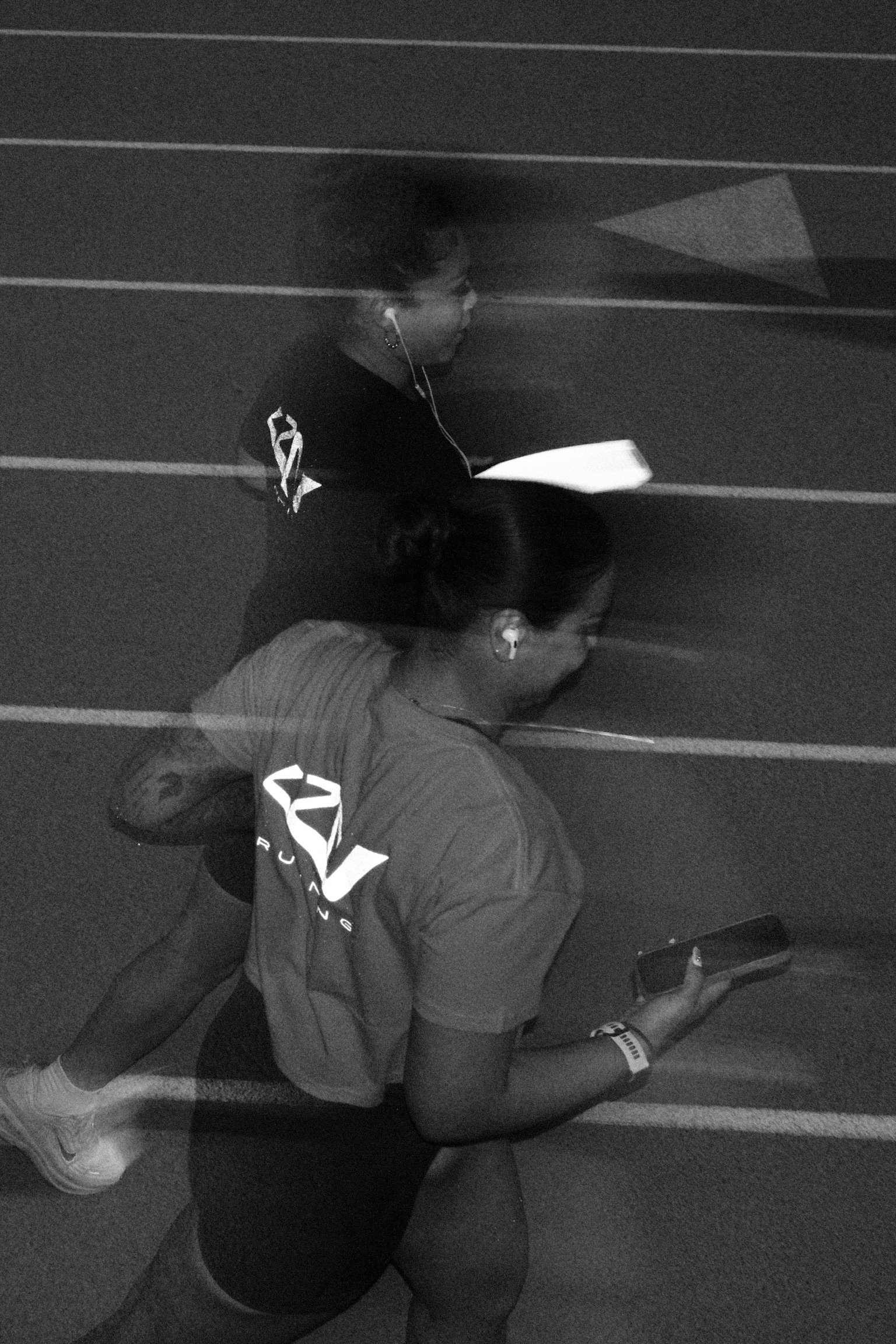 Two runners, one male and one female, running on a track, both wearing sports shirts with a logo, and wearing earphones.