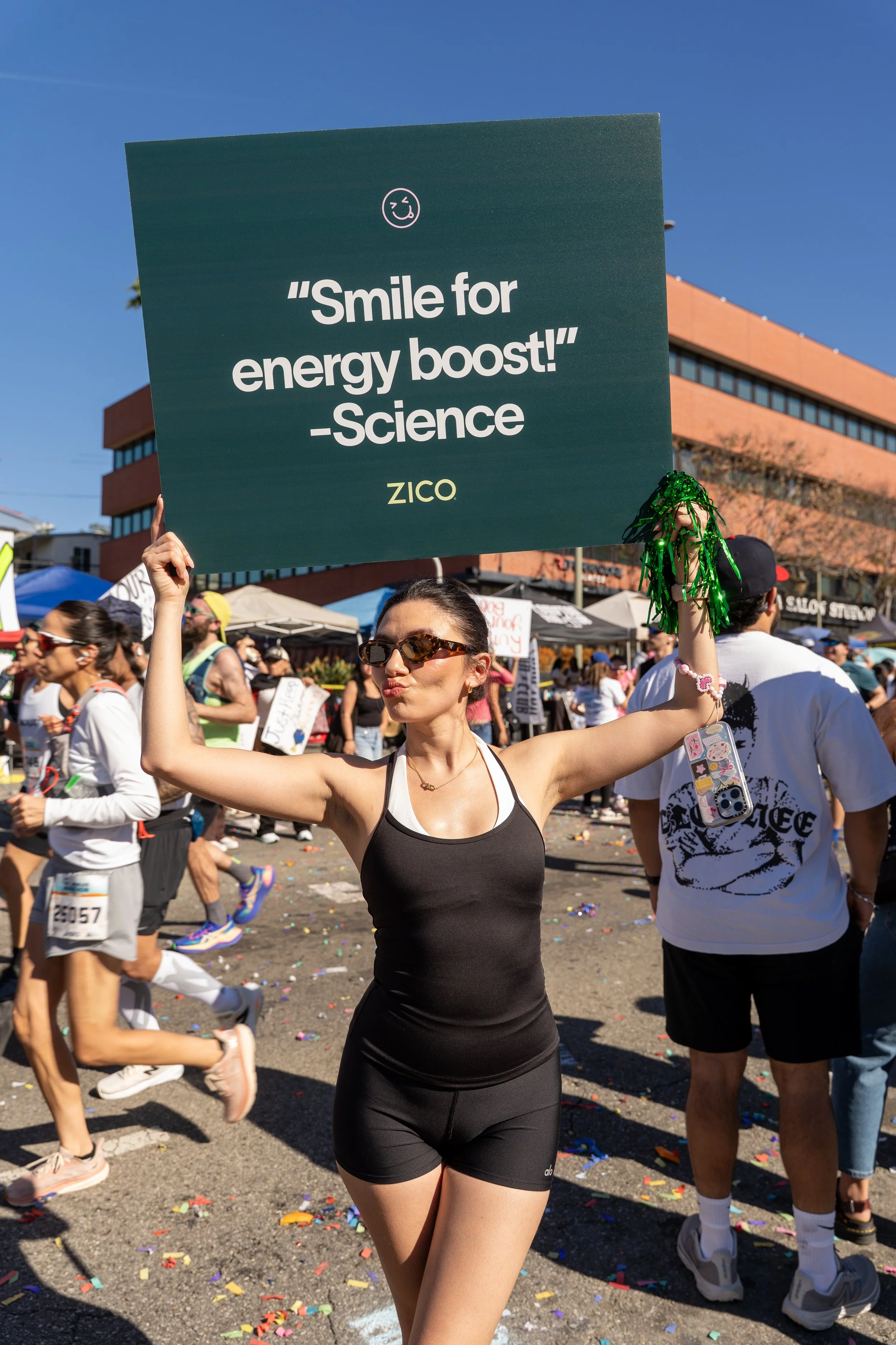A woman in black athletic attire and sunglasses holding a sign that says 'Smile for energy boost! -Science ZICO' at a lively outdoor event with other participants and colorful tents.