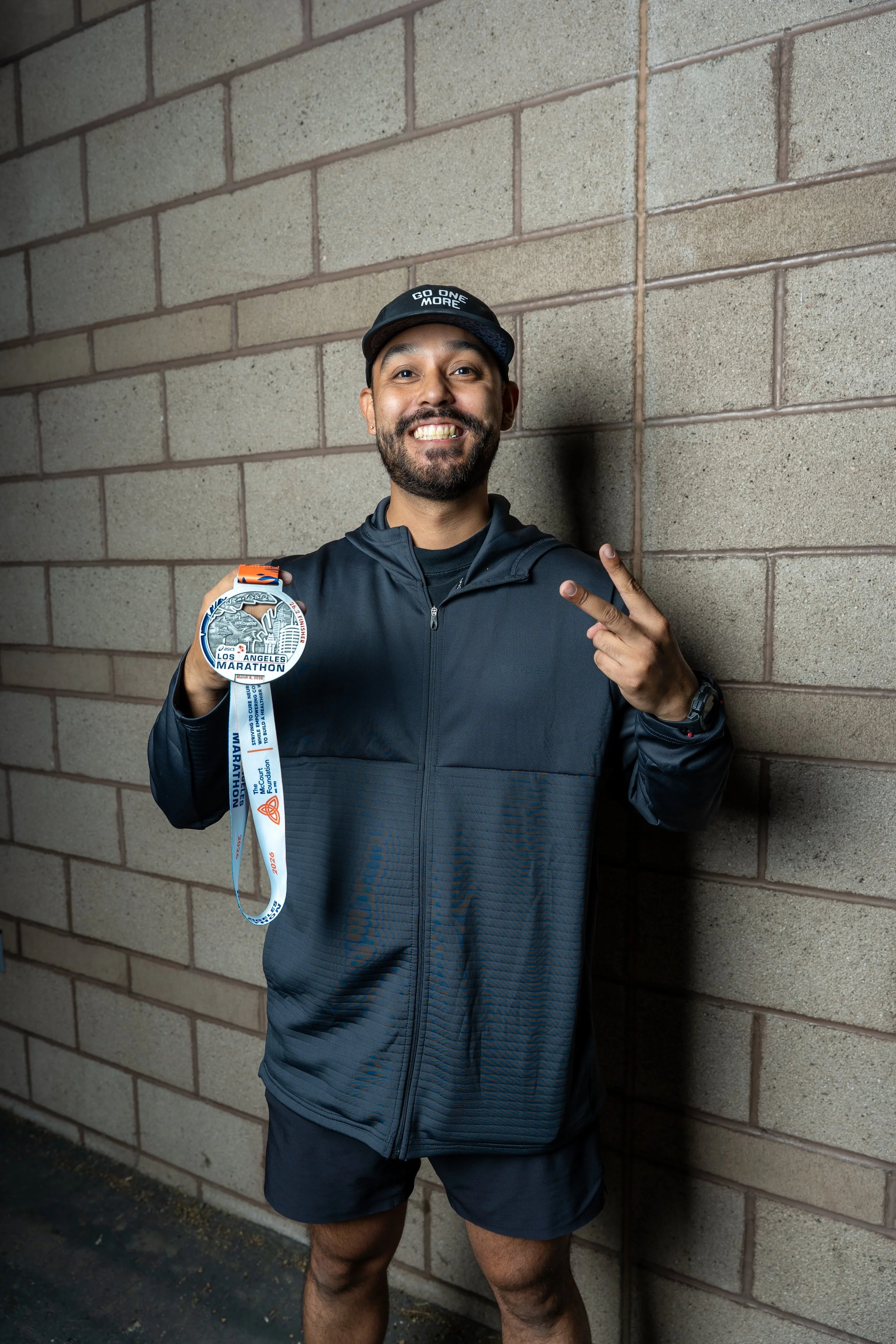 Man smiling and pointing at his marathon medal after a race, standing against a brick wall.