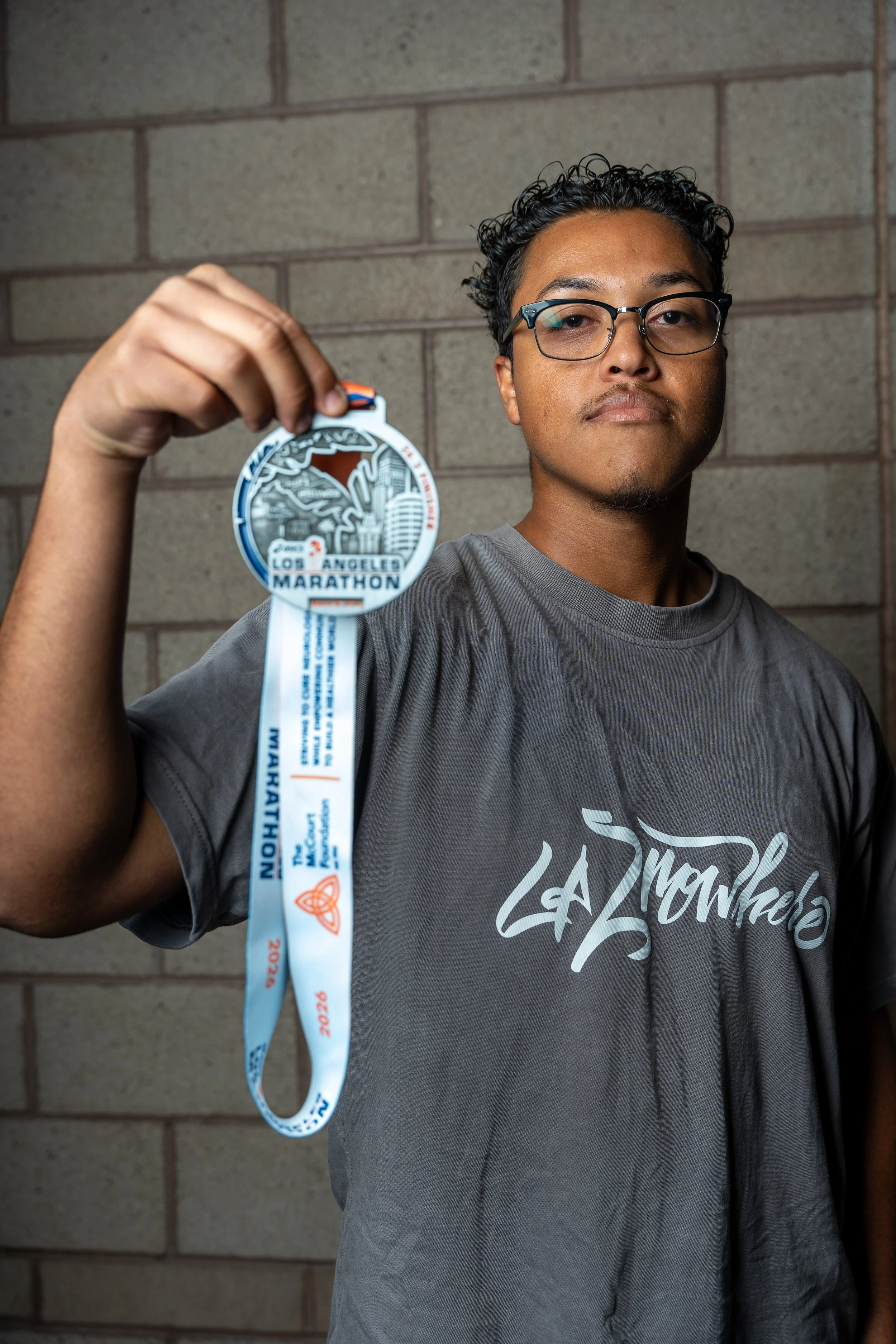 A young man wearing glasses and a gray La Mohale t-shirt holding a Los Angeles Marathon medal in front of a brick wall.