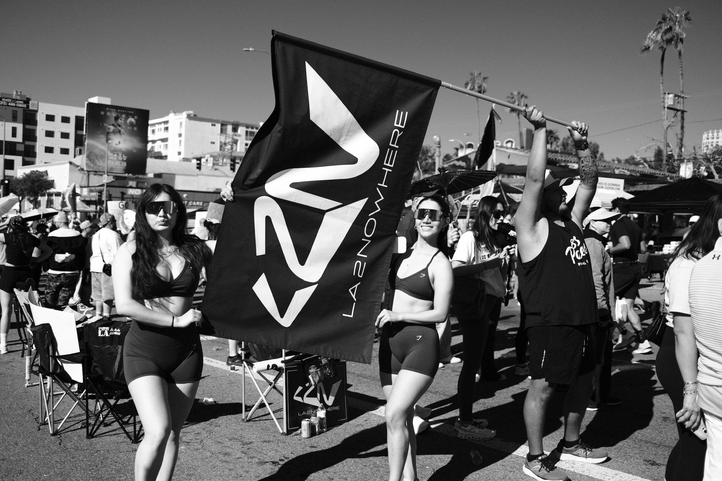 People participating in an outdoor event, some holding a large flag with a logo that reads 'LA 2 NOW HERE', with tents and buildings in the background.
