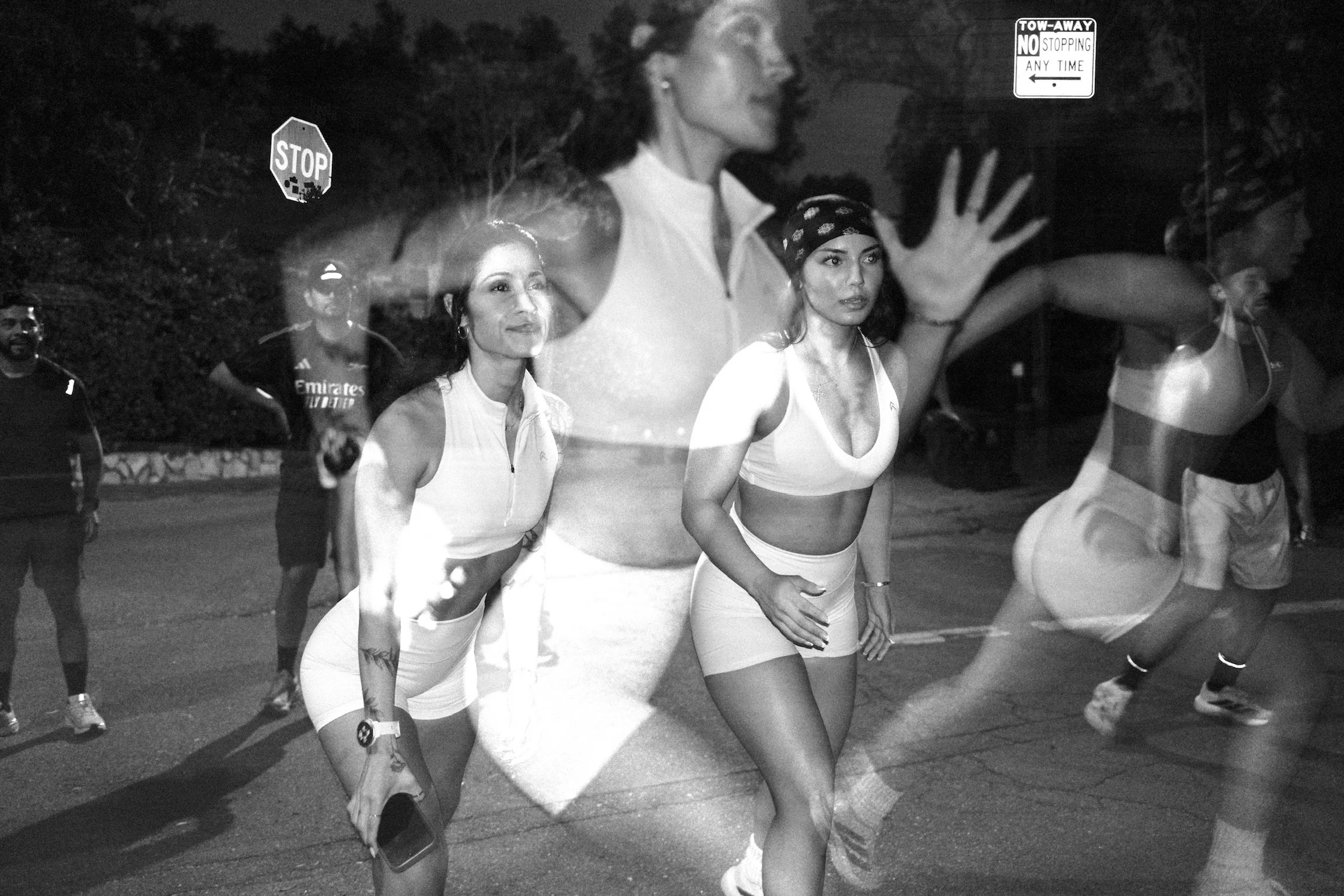 A black and white photo of women running on the street at night, with a painted crosswalk and stop sign visible, and a sign indicating no stopping at any time.