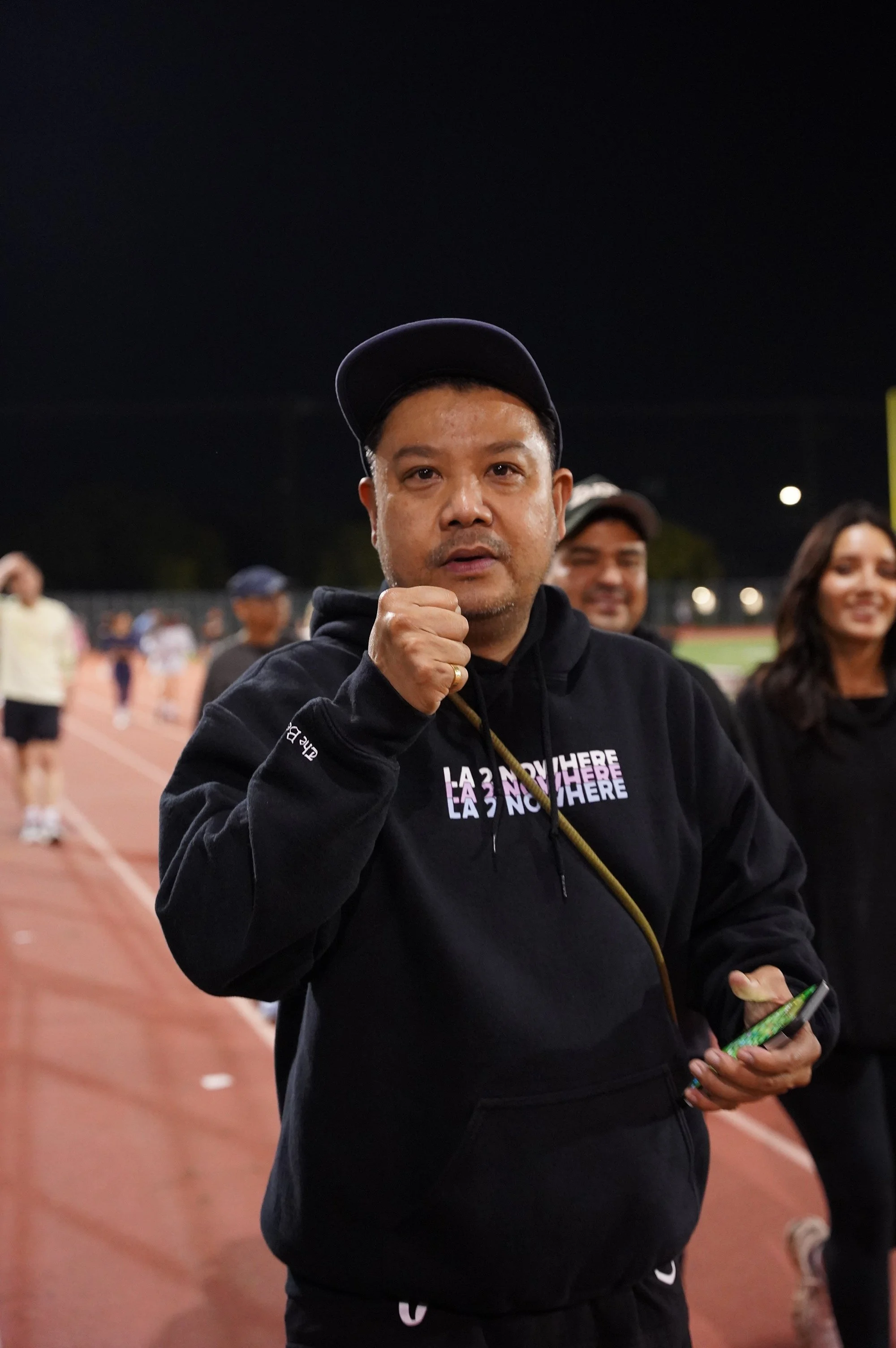 A man on a running track at night makes a fist pump gesture, wearing a black hoodie and cap, with others in the background at a sports event.