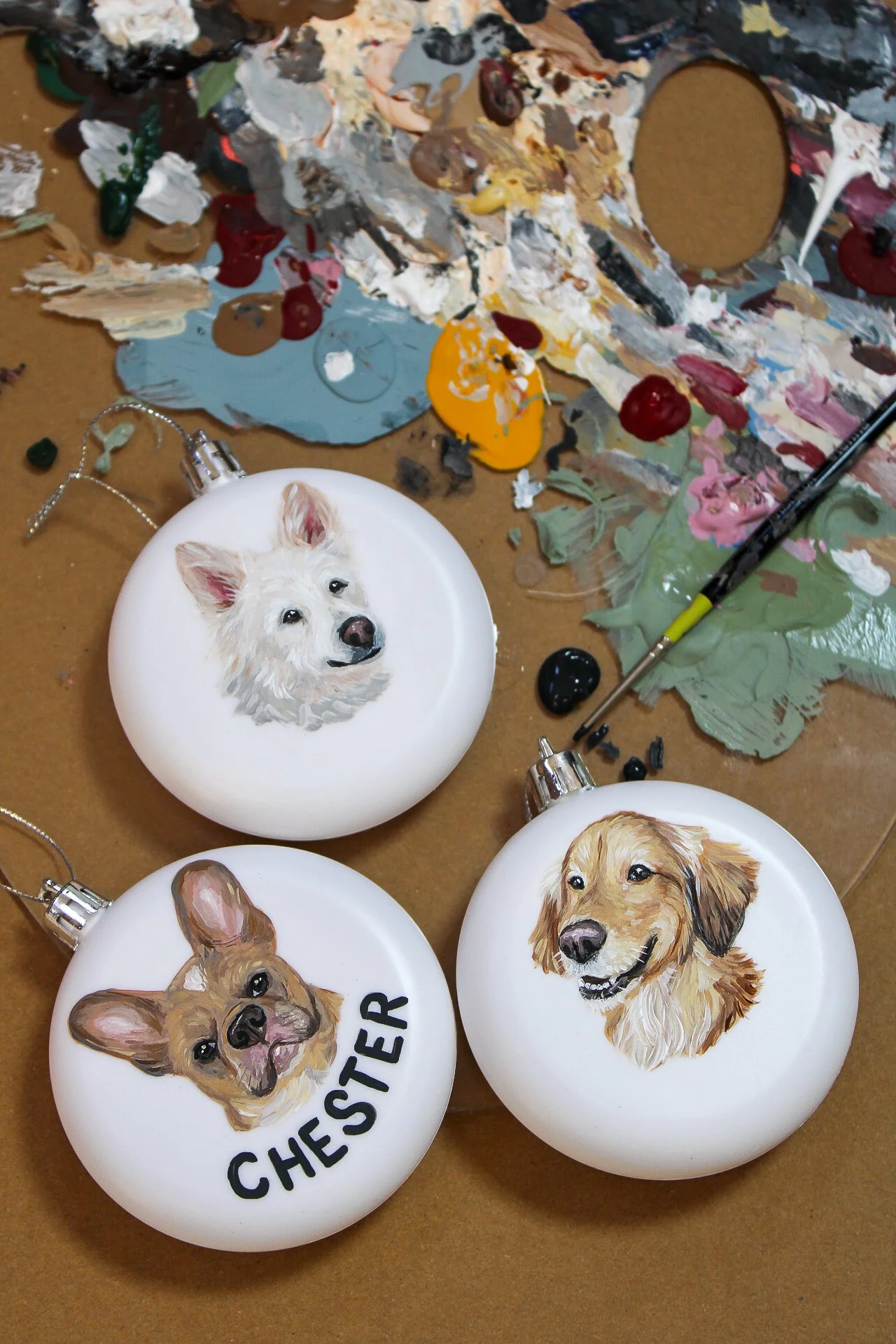 Decorative ornaments featuring hand-painted portraits of different dogs, placed on a work surface with paint smudges and tools around.