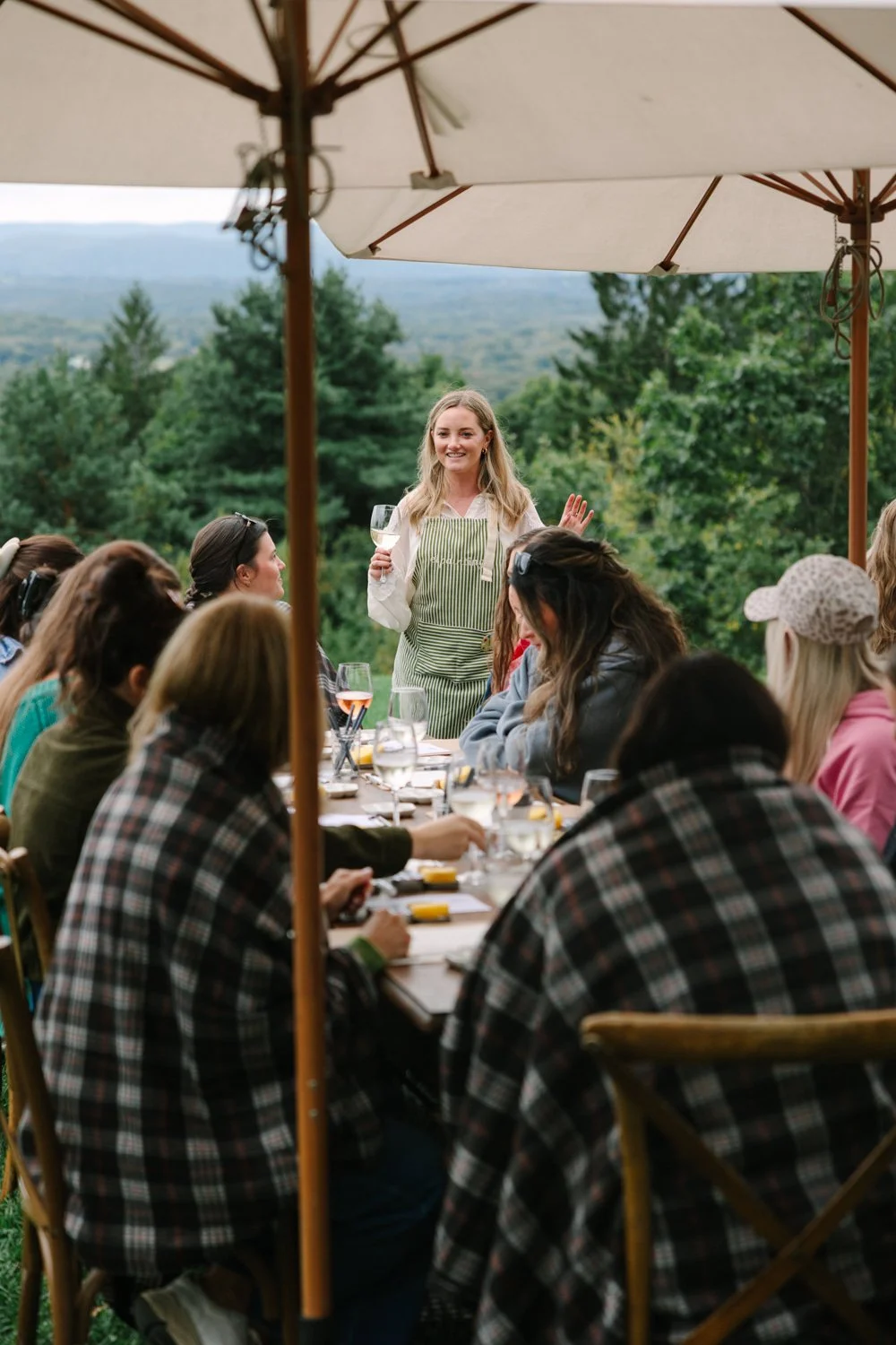 A woman standing and speaking to a group of people seated outdoors at a long table with umbrellas, surrounded by trees and mountains in the background.