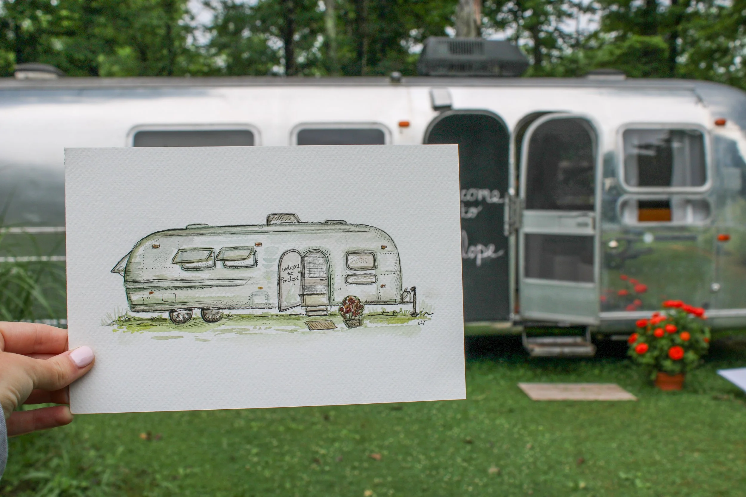 A person holding a watercolor drawing of a camper trailer in front of a real camper trailer, with trees and flowers in the background.