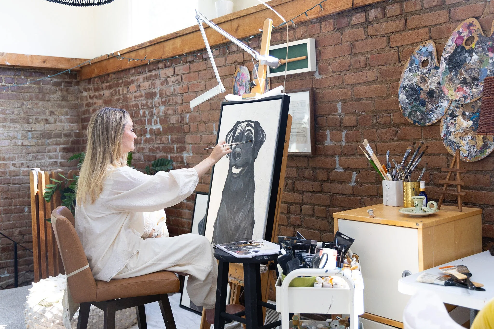 A woman with long blonde hair wearing a white dress is sitting and painting a portrait of a dog on a vertical canvas in an art studio with an exposed brick wall. The studio has art supplies, brushes, and canvases on the wall and table, and a small plant is visible near her.