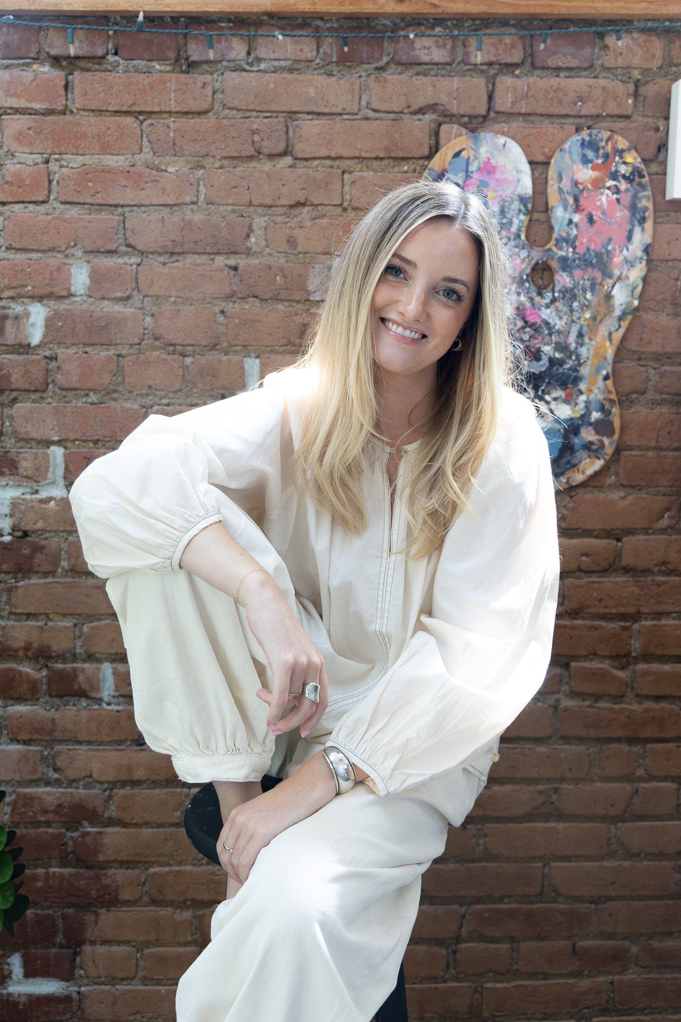A woman with long blonde hair smiling while sitting on a black chair in front of a brick wall, with abstract paintings in the background.