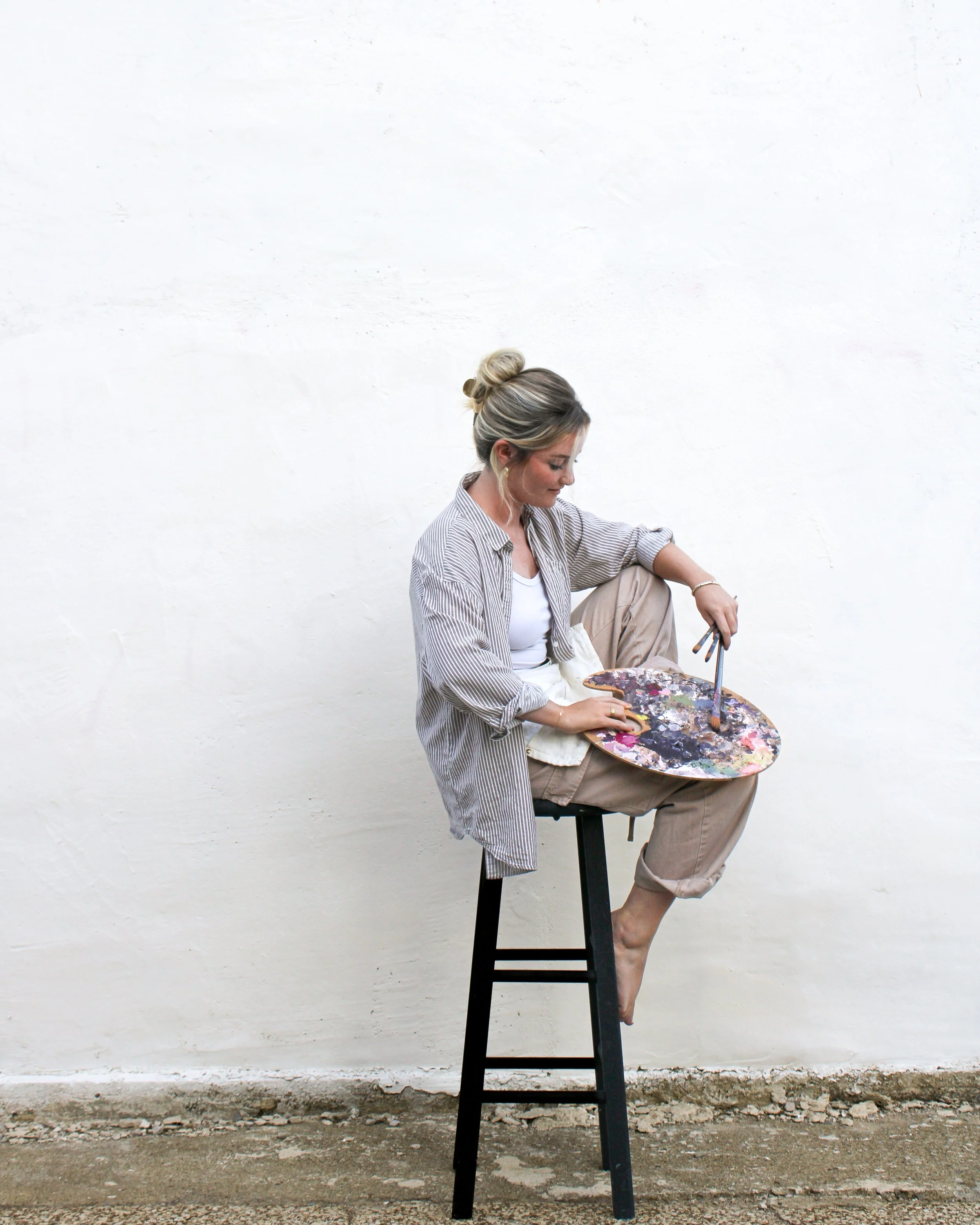 A woman with blonde hair tied in a bun sitting on a tall black stool, painting on an oval canvas with a large paintbrush in her right hand. She is wearing a striped shirt and beige pants, against a plain white wall background.