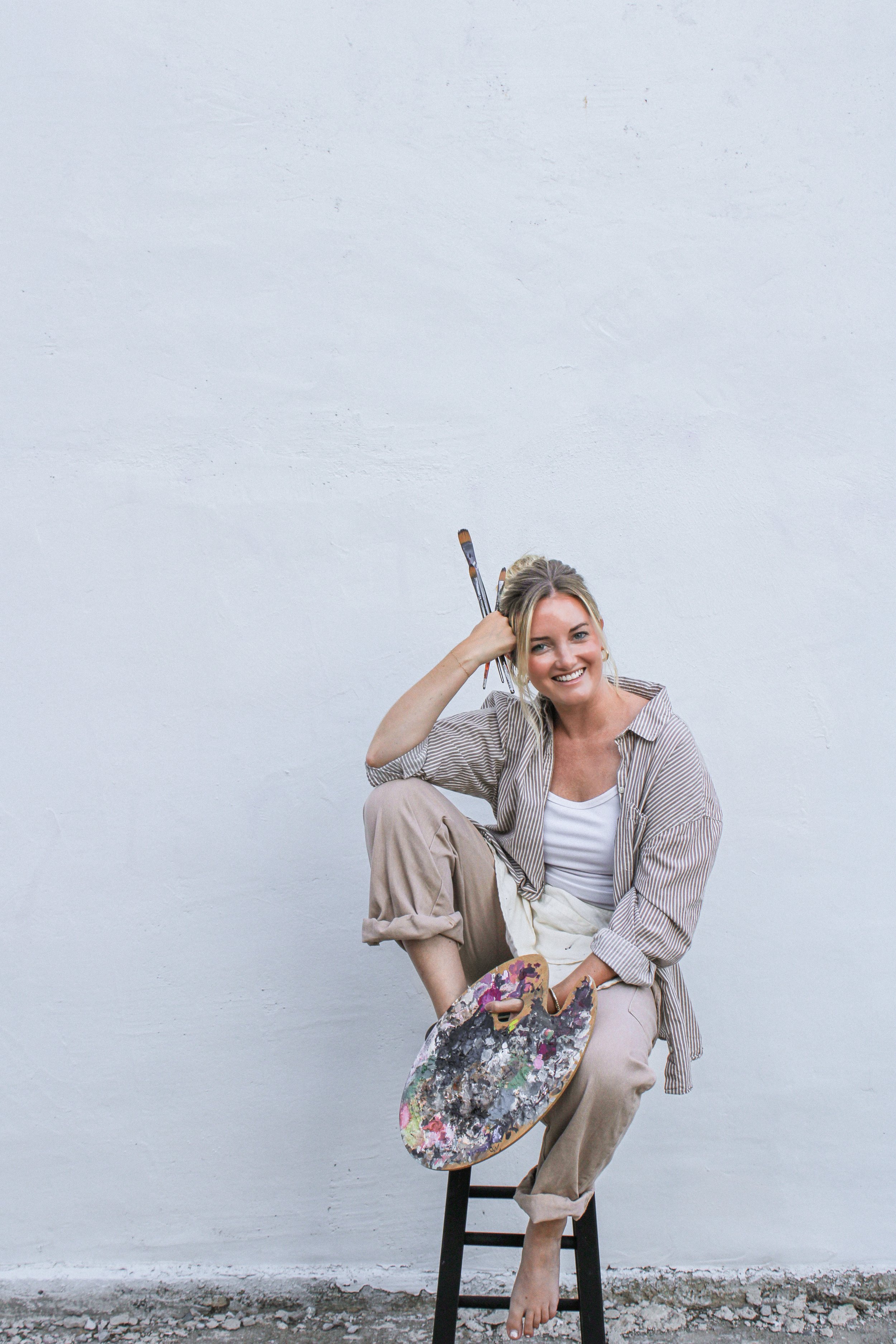 Young woman sitting on a stool, holding a paint palette, with paintbrushes in her hair, smiling, against a plain white wall.