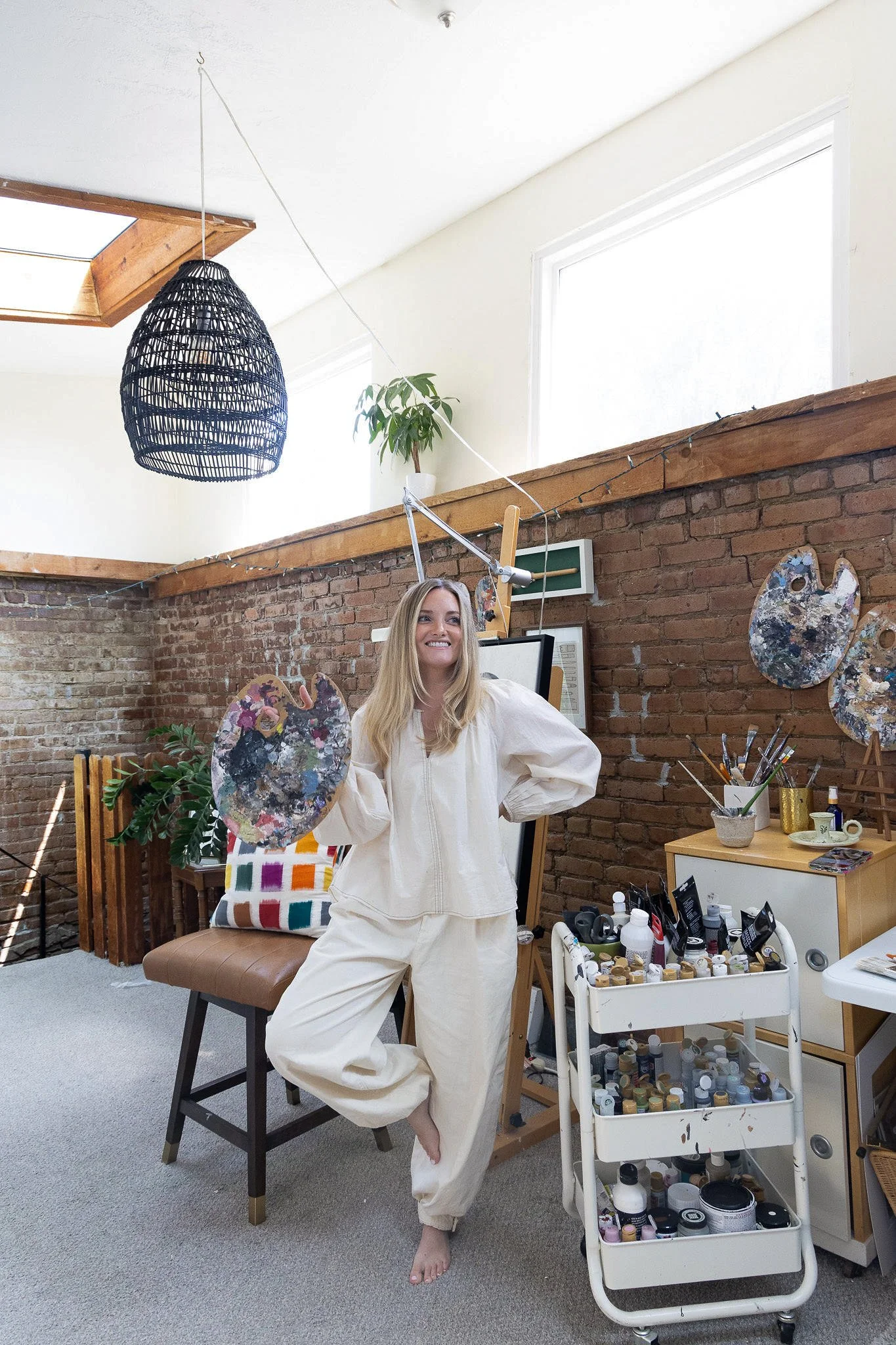 A woman standing in an art studio, holding a paint palette, with a large cart filled with paints and art supplies nearby. The room has a brick wall, two windows, and various art pieces and tools, with natural light coming in.