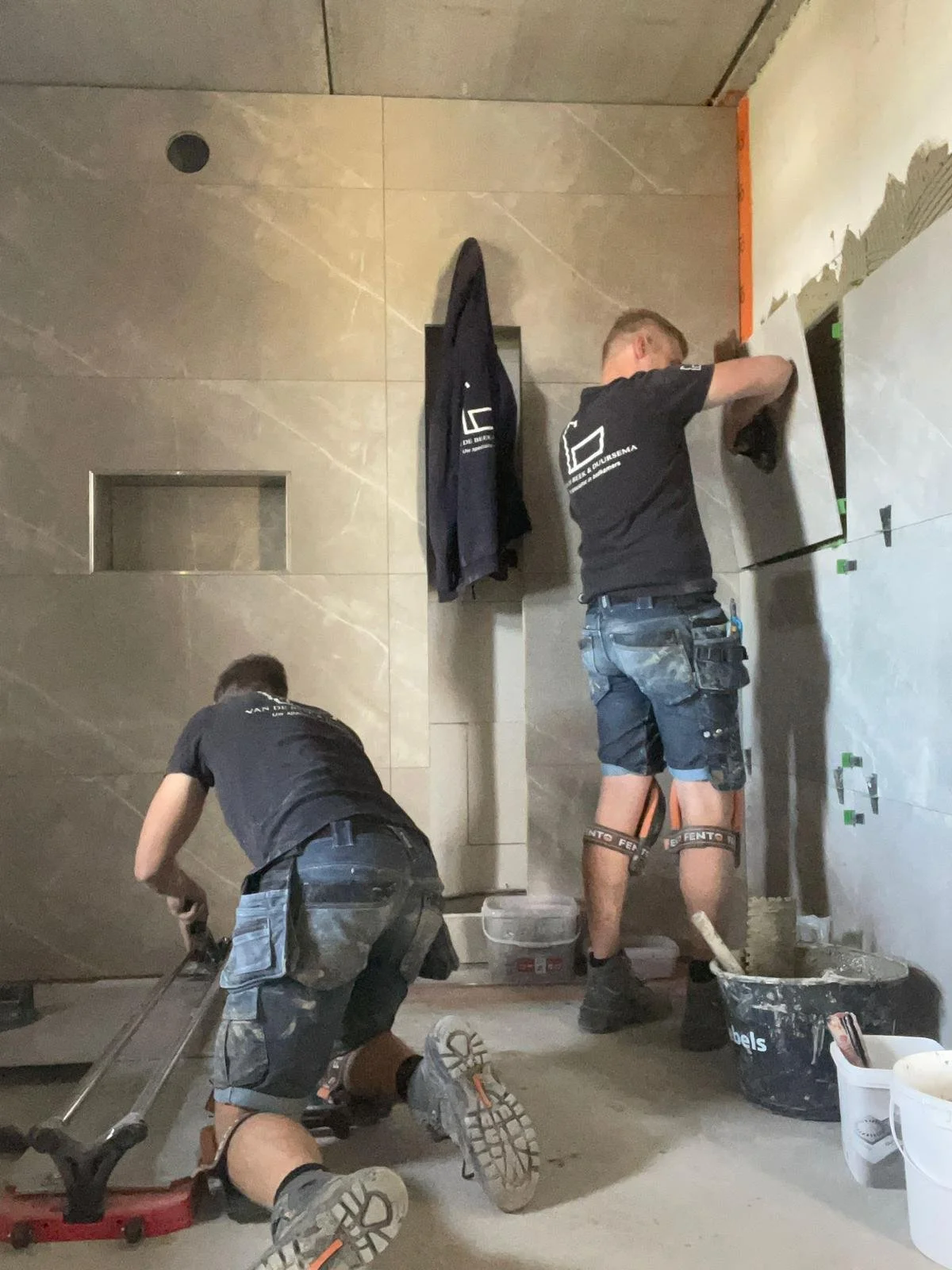 Twee mannen werken in een kamer, een ligt op de vloer en werkt met een gereedschap, de andere staat en plakt tegels aan de muur.