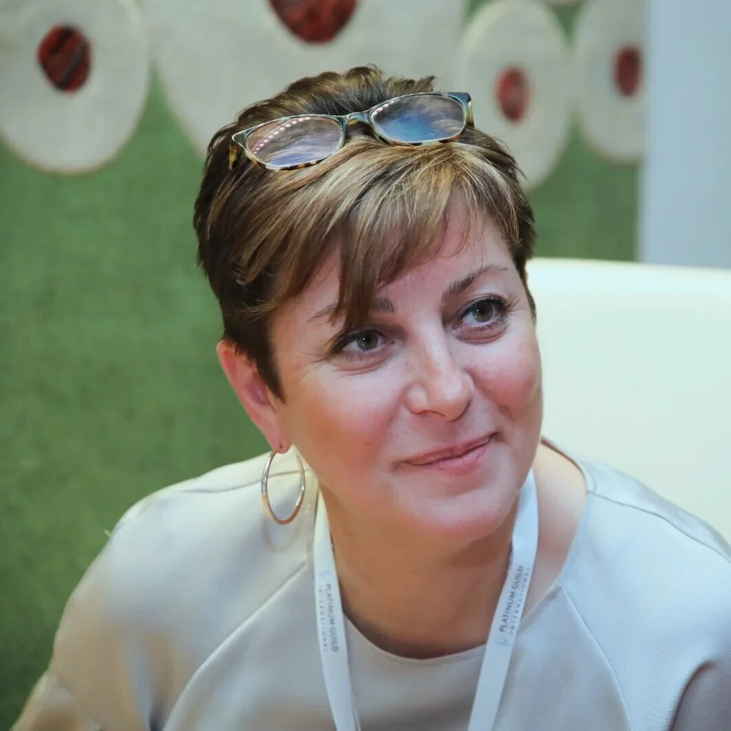 A woman with short brown hair, wearing hoop earrings and glasses on her head, sitting indoors with a green background featuring fruit images.