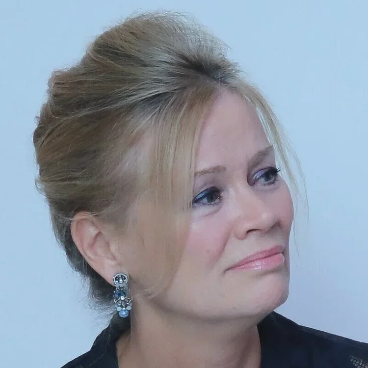 A woman with blonde hair styled in an elegant updo, wearing earrings and a dark top, looking slightly to her left with a neutral expression.