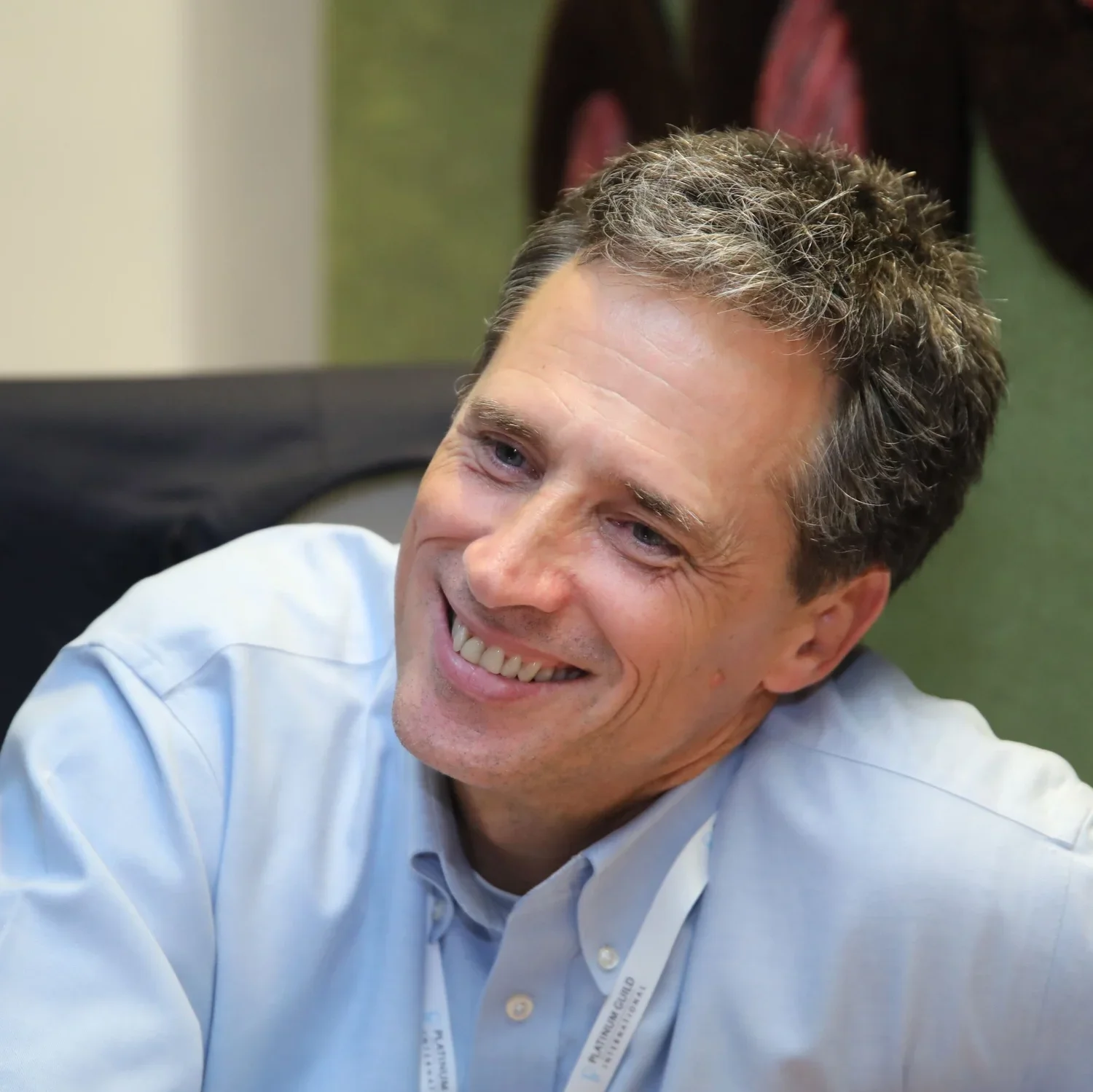 A man with brown, slightly curly hair smiling and looking to his left. He is wearing a light blue collared shirt, seated indoors with blurred background.