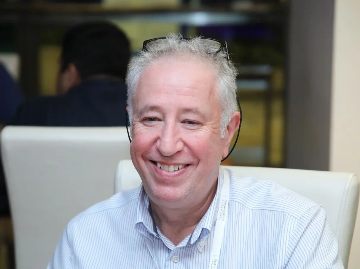 A smiling middle-aged man with gray hair and glasses on his head wearing a light blue striped collared shirt, sitting in an office setting.