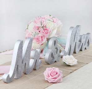 Silver glittered 'Mr & Mrs' sign with pink and white roses on a beige table with a pink ribbon.