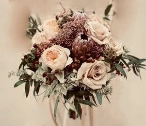 A bouquet of cream and blush roses, purple flowers, and greenery in a neutral-toned vase.