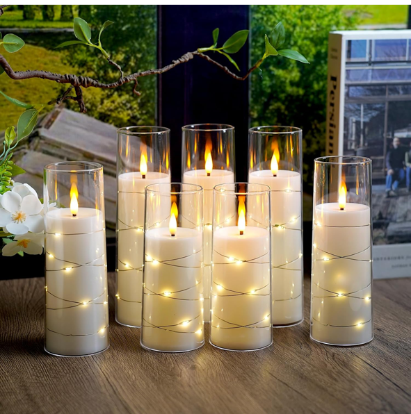 Seven tall glass hurricanes with white candles and string lights inside, arranged on a wooden surface with a plant and window in the background.