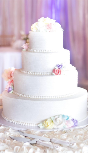 A four-tiered white wedding cake decorated with pastel-colored flowers, set on a table with flower petals and cake servers.